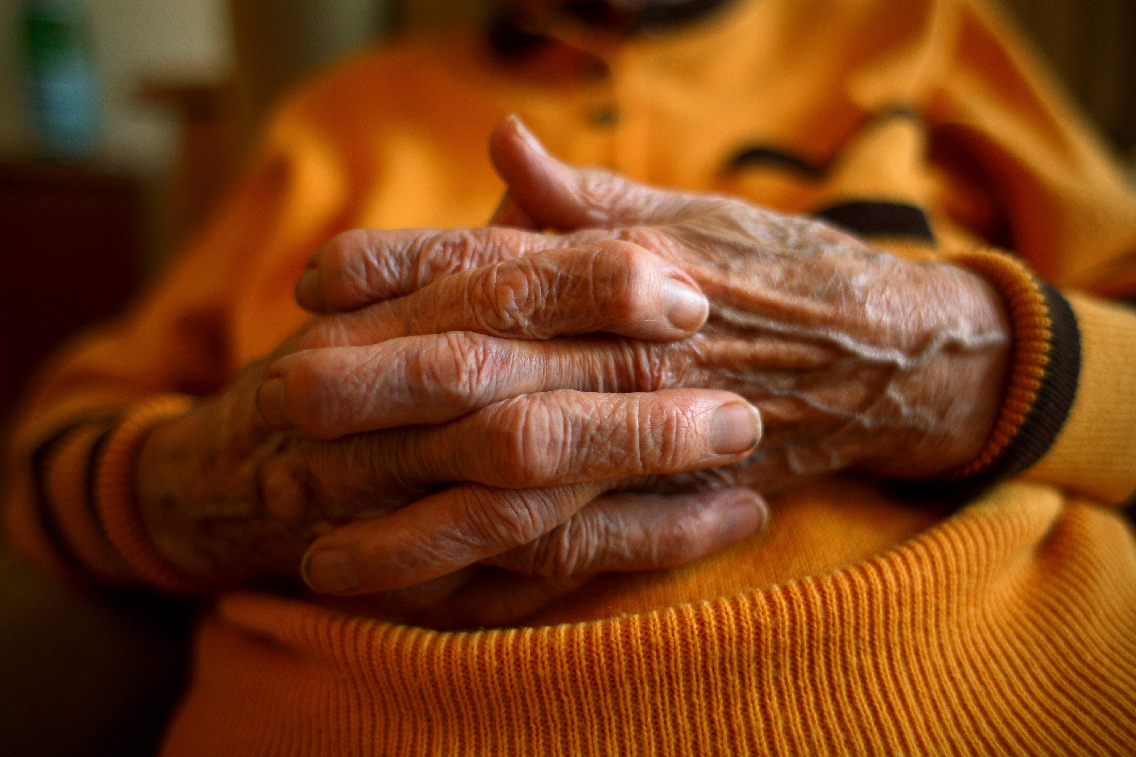Photo of an elderly person’s hands clasped together, wearing an orange jumper, with focus on the wrinkles and texture.