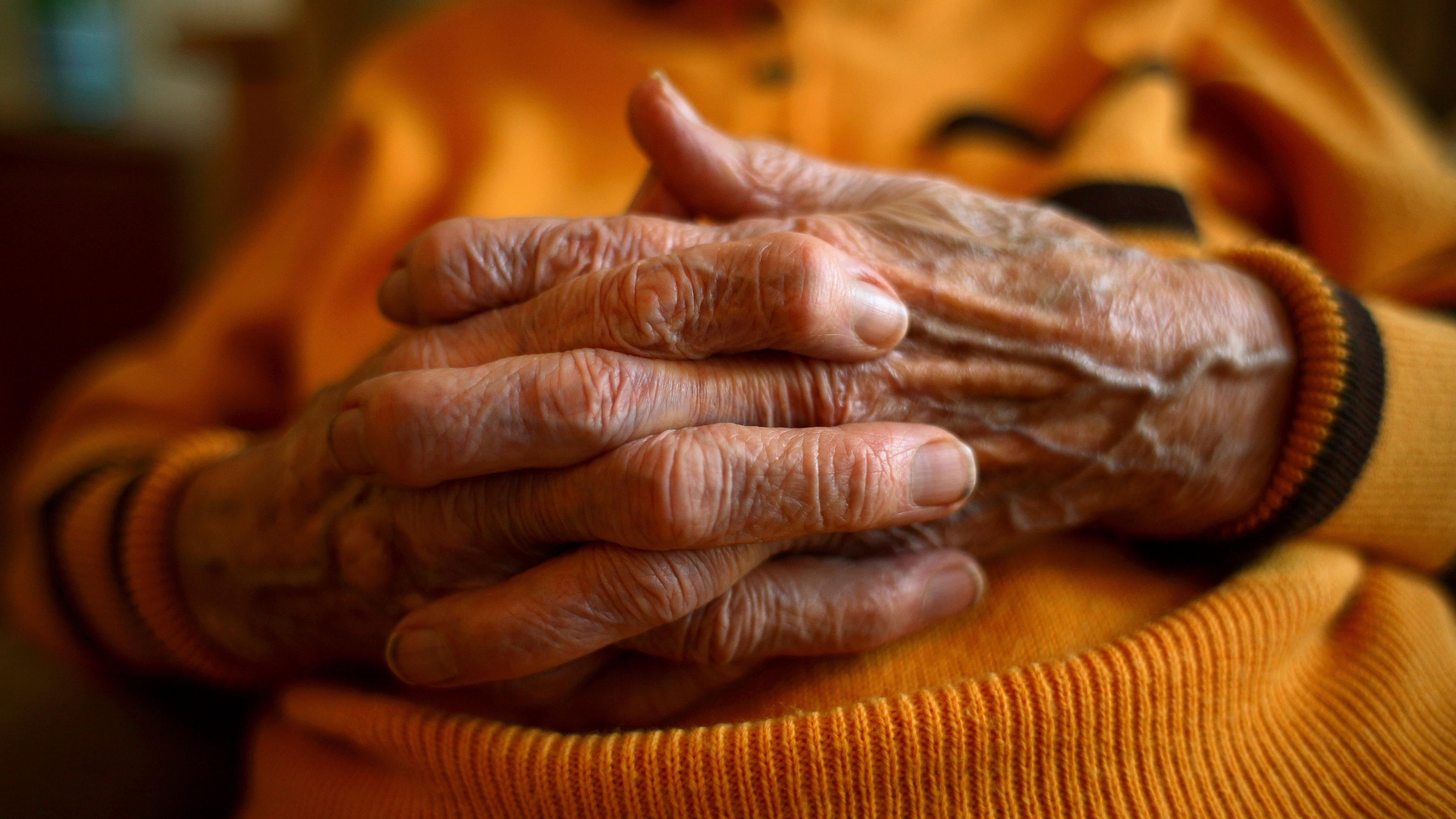 Photo of an elderly person’s hands clasped together, wearing an orange jumper, with focus on the wrinkles and texture.