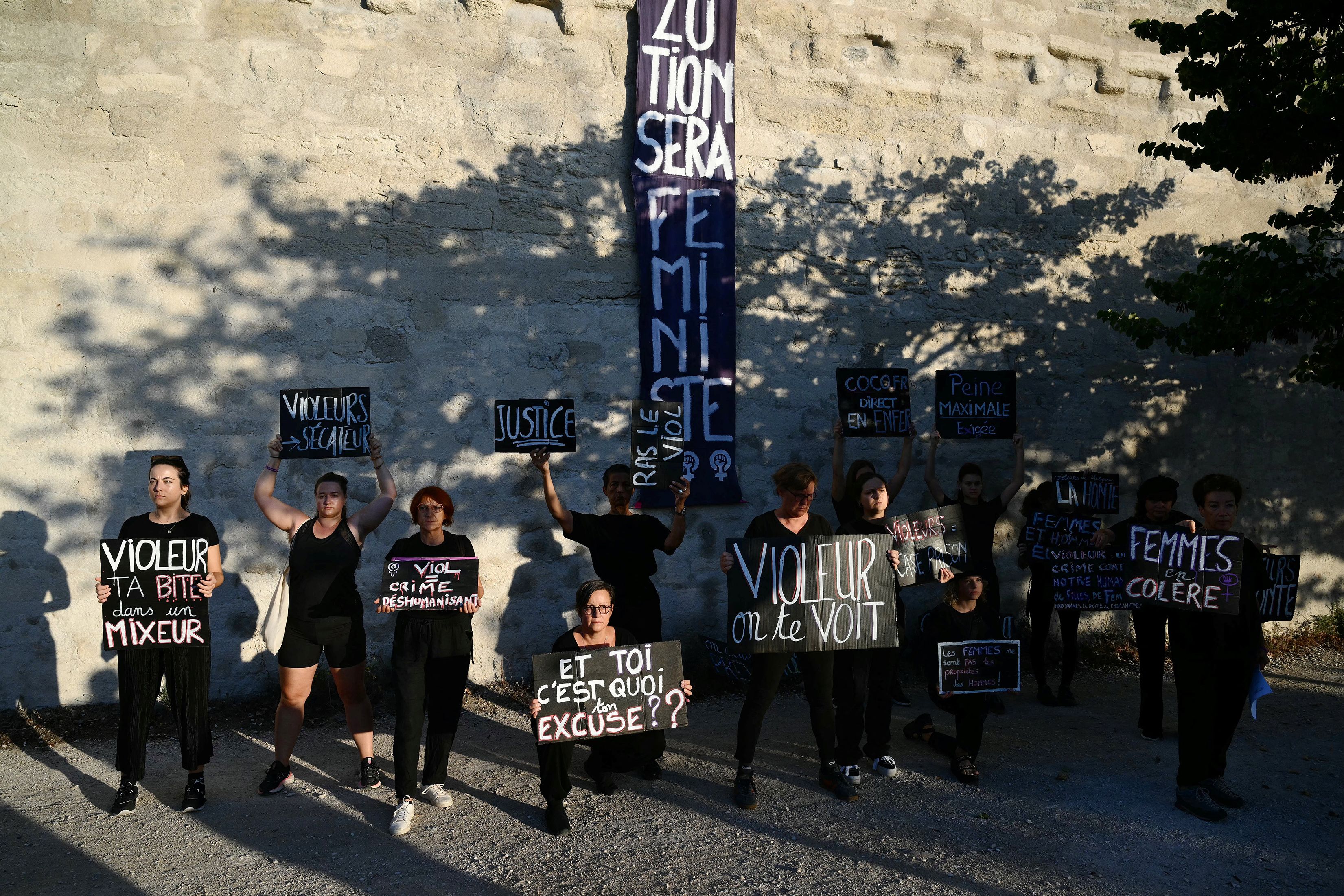 People holding protest signs in French against a stone wall, highlighting feminist and anti-violence messages.