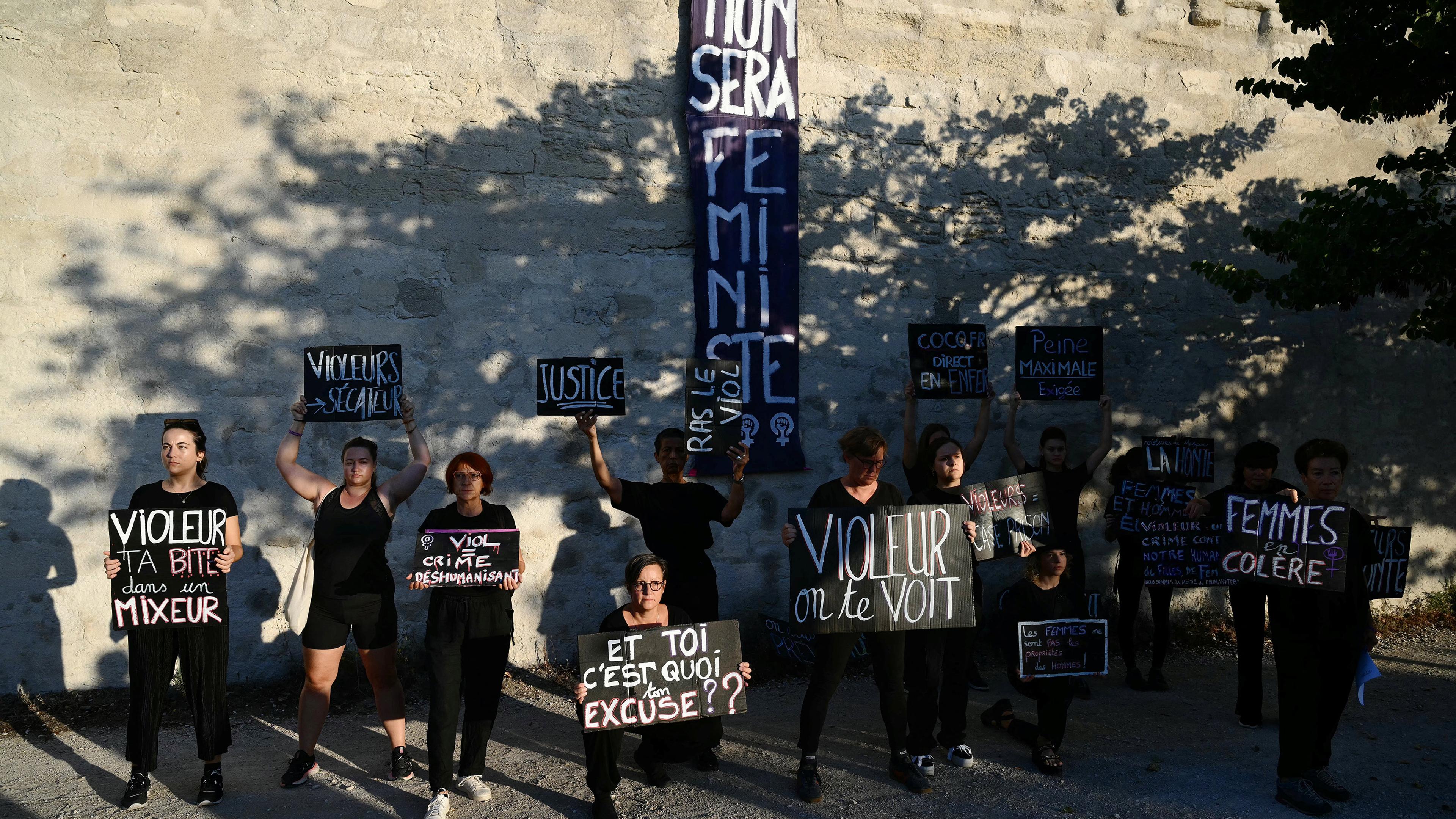 People holding protest signs in French against a stone wall, highlighting feminist and anti-violence messages.