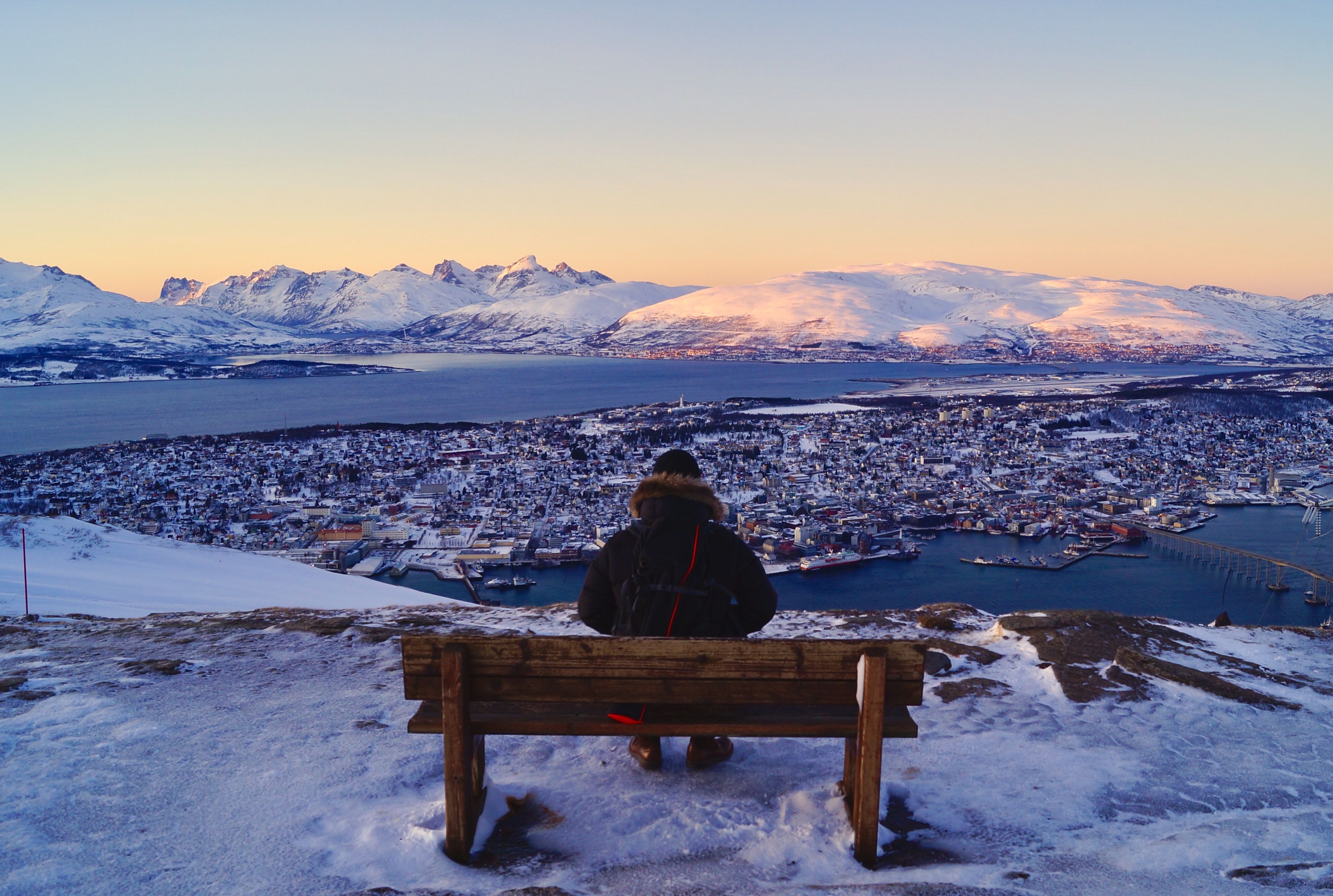 Photo of a person on a bench overlooking a snowy town, fjord and mountains at sunset.