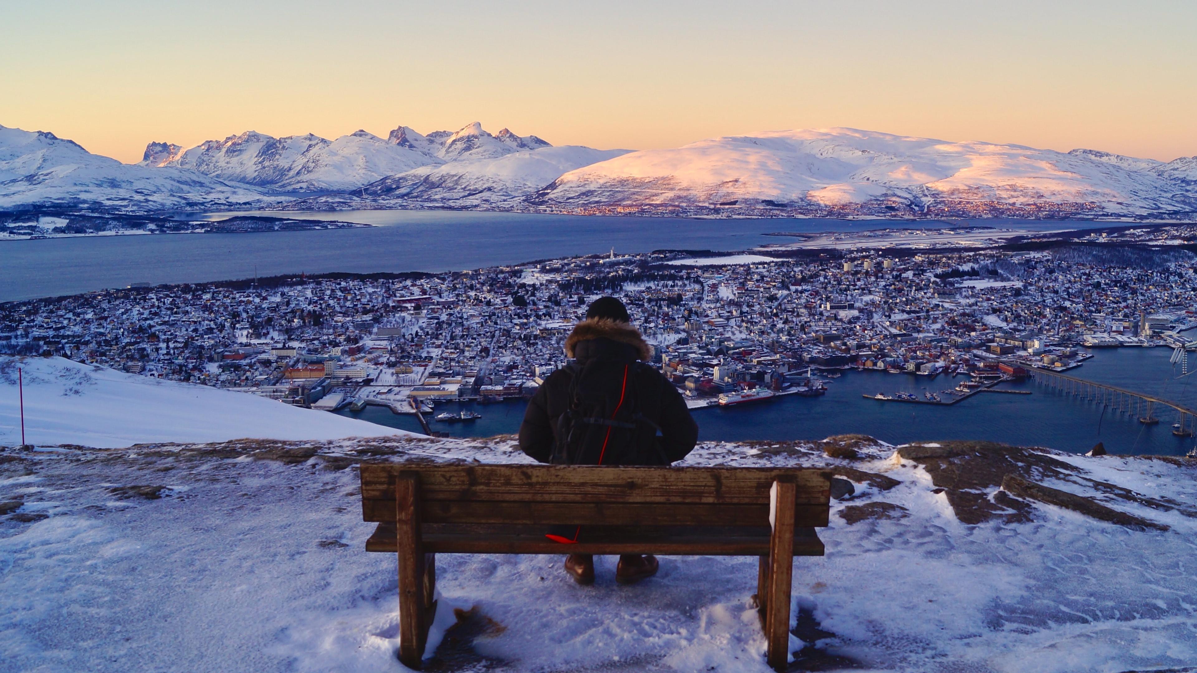 Photo of a person on a bench overlooking a snowy town, fjord and mountains at sunset.