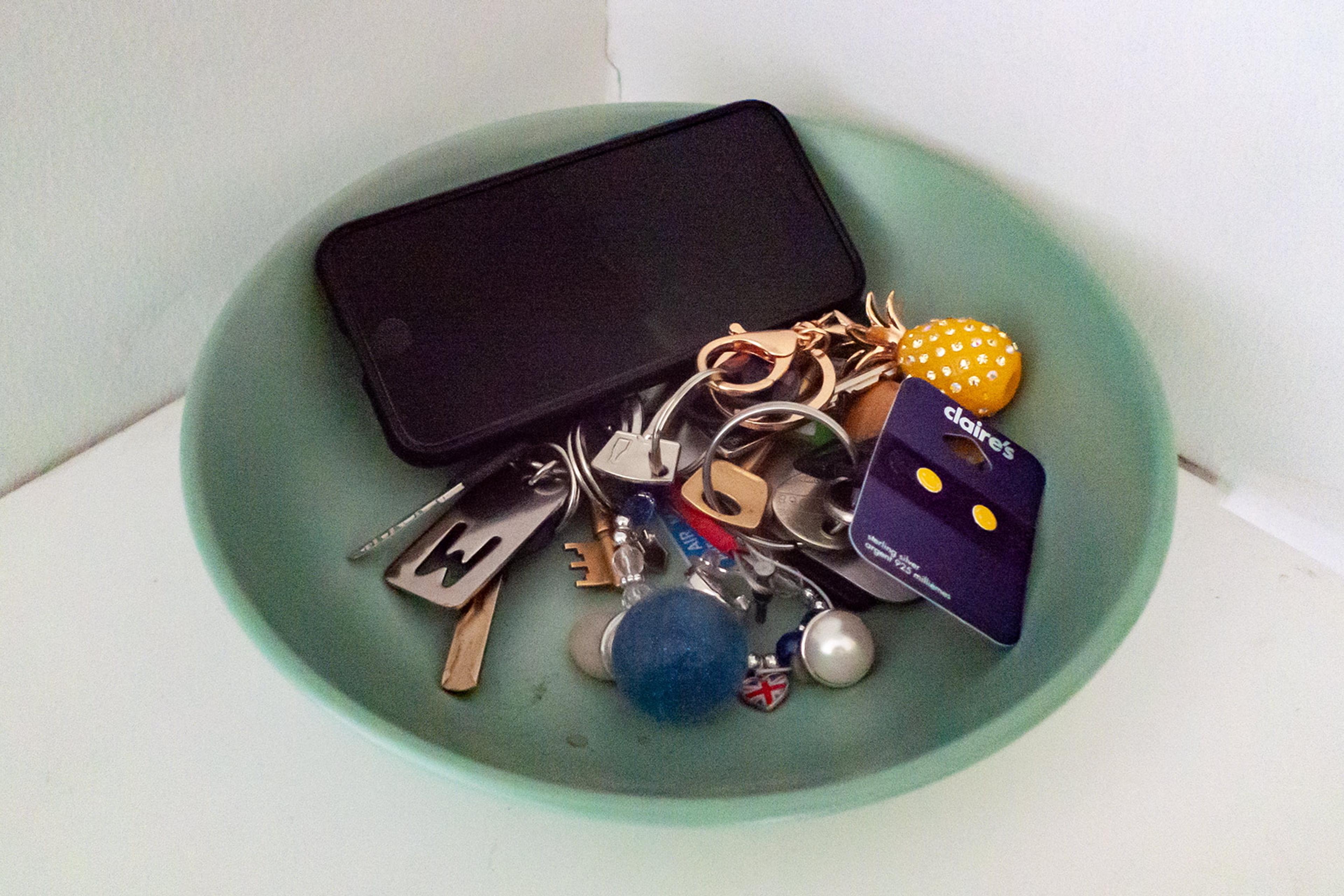 Photo of a green bowl holding a smartphone various keys a yellow accessory and a blue card on a white surface.