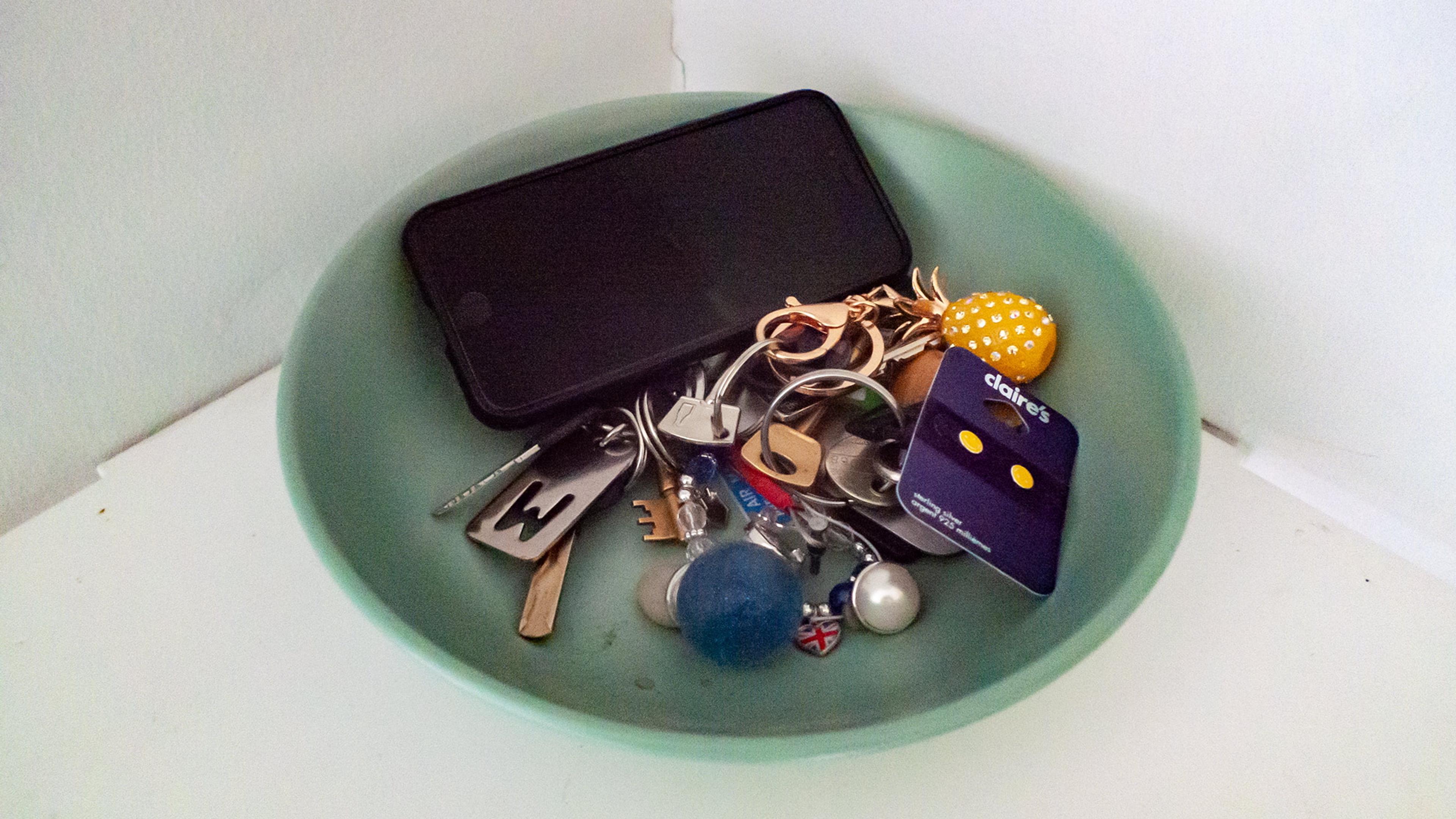 Photo of a green bowl holding a smartphone various keys a yellow accessory and a blue card on a white surface.