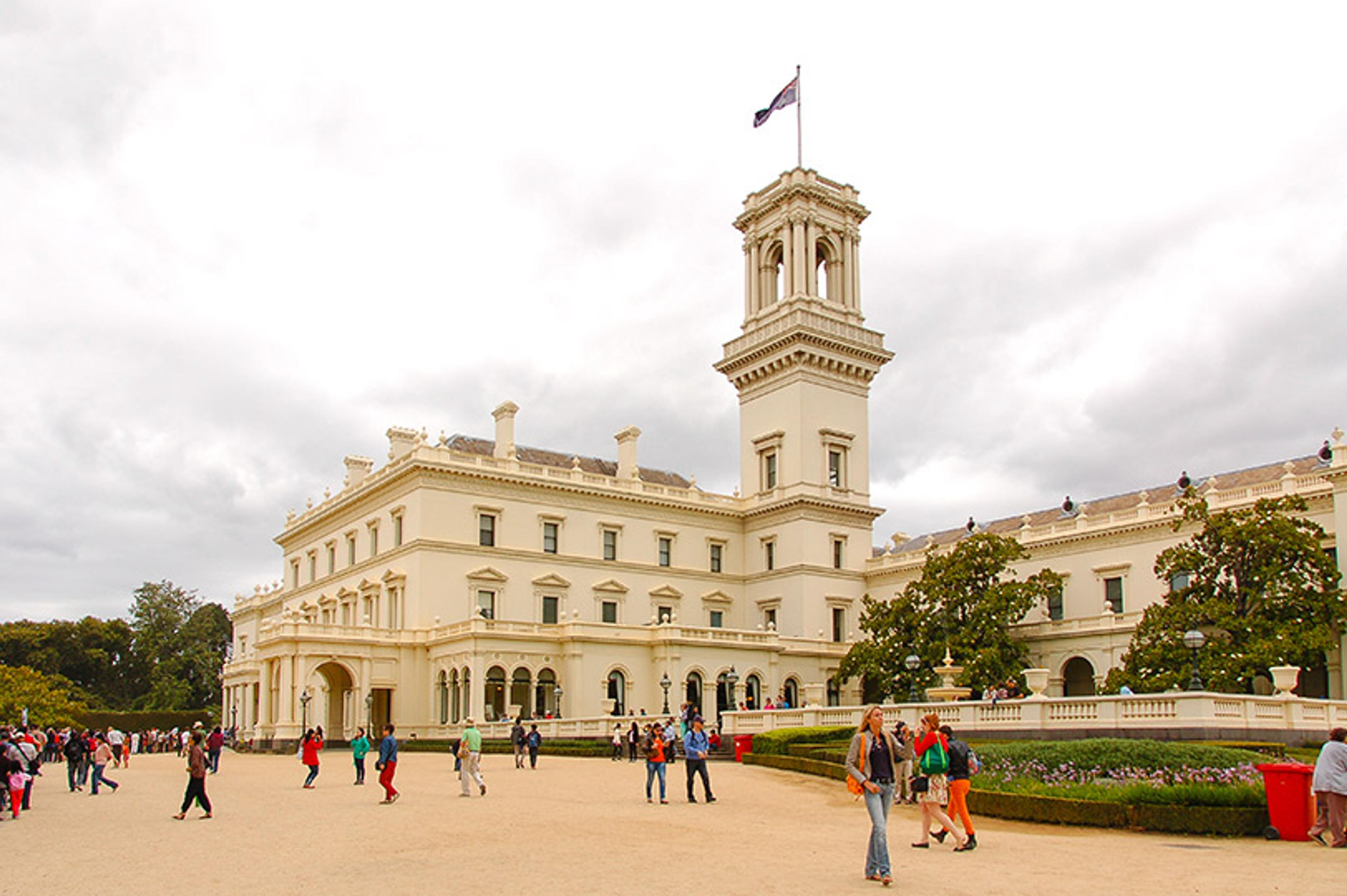 A grand white building with a tall tower, surrounded by people walking on a pathway on a cloudy day.