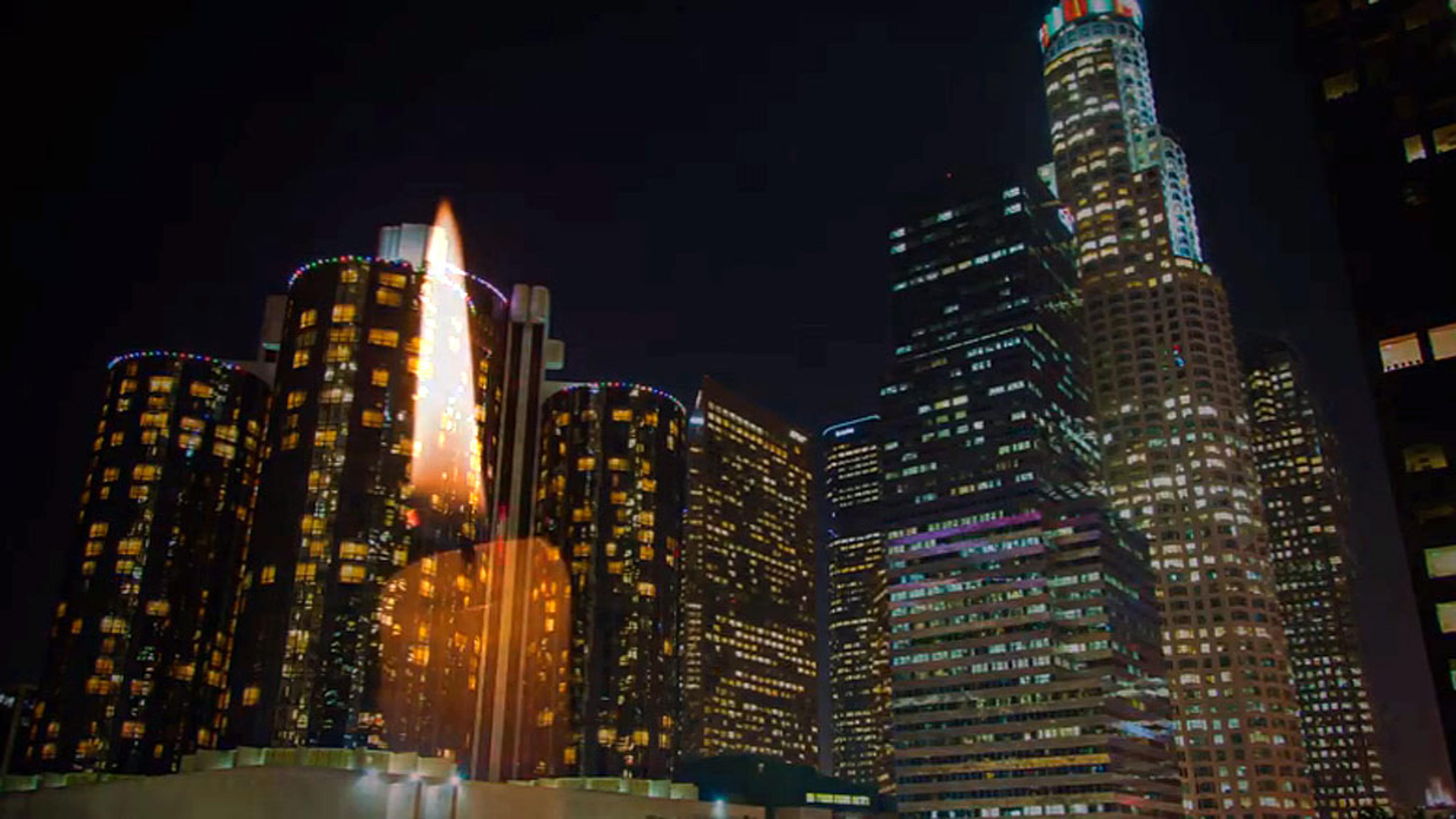 Illuminated skyscrapers at night in a city, with a bright, blurred object resembling a candle flame in the foreground.