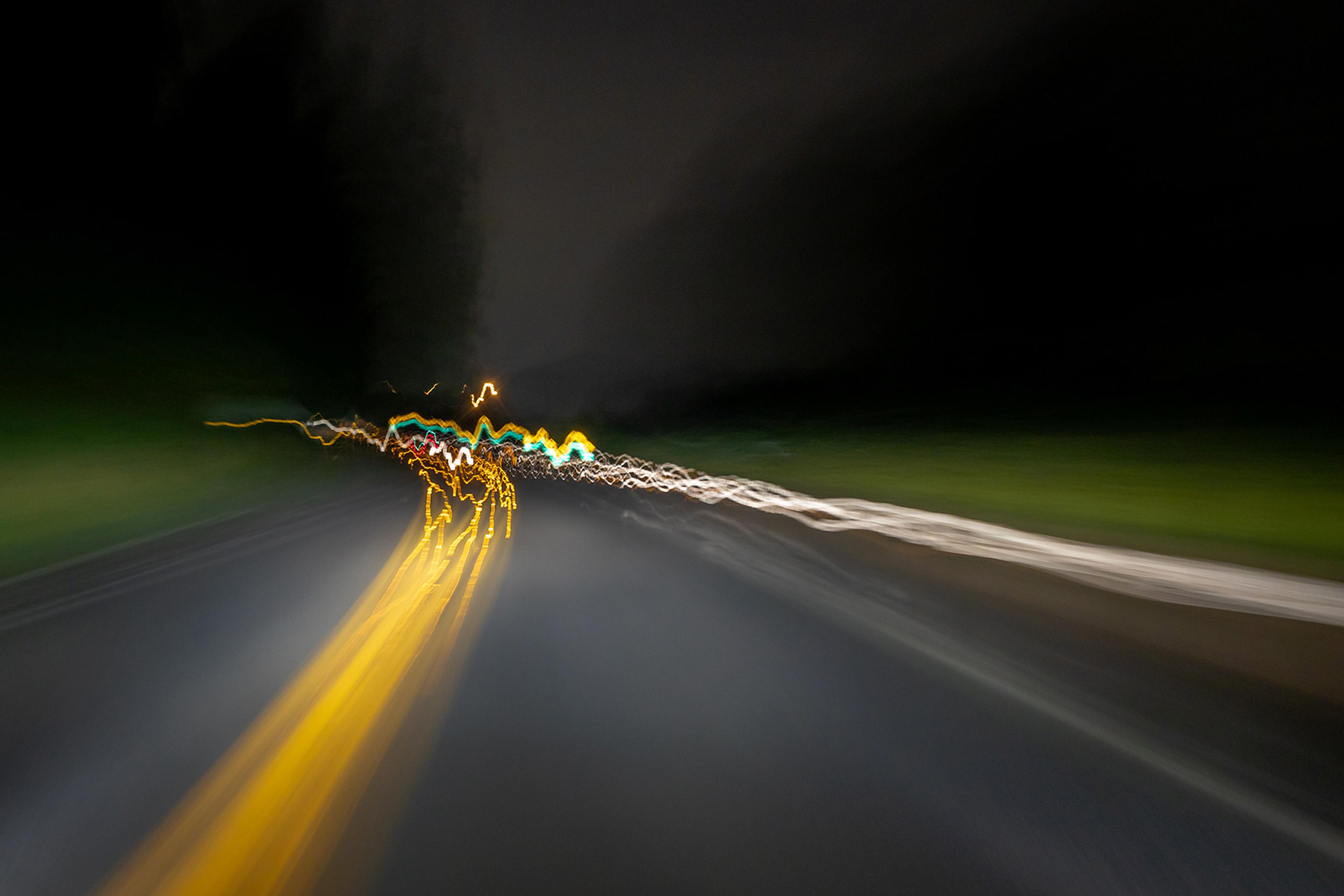 Long-exposure photo of a road at night with blurry, colourful light trails from car headlights and taillights against a dark background.