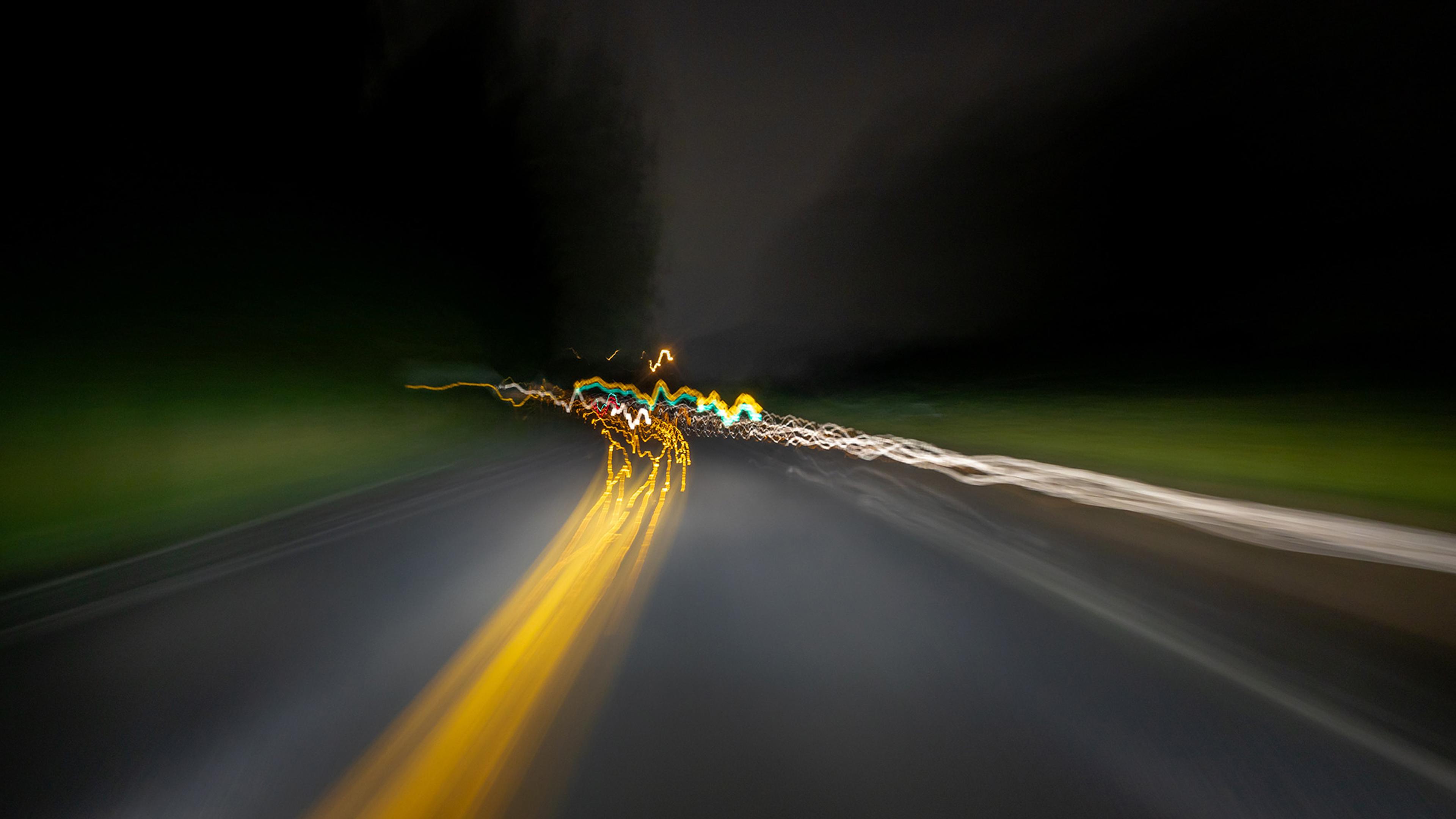 Long-exposure photo of a road at night with blurry, colourful light trails from car headlights and taillights against a dark background.