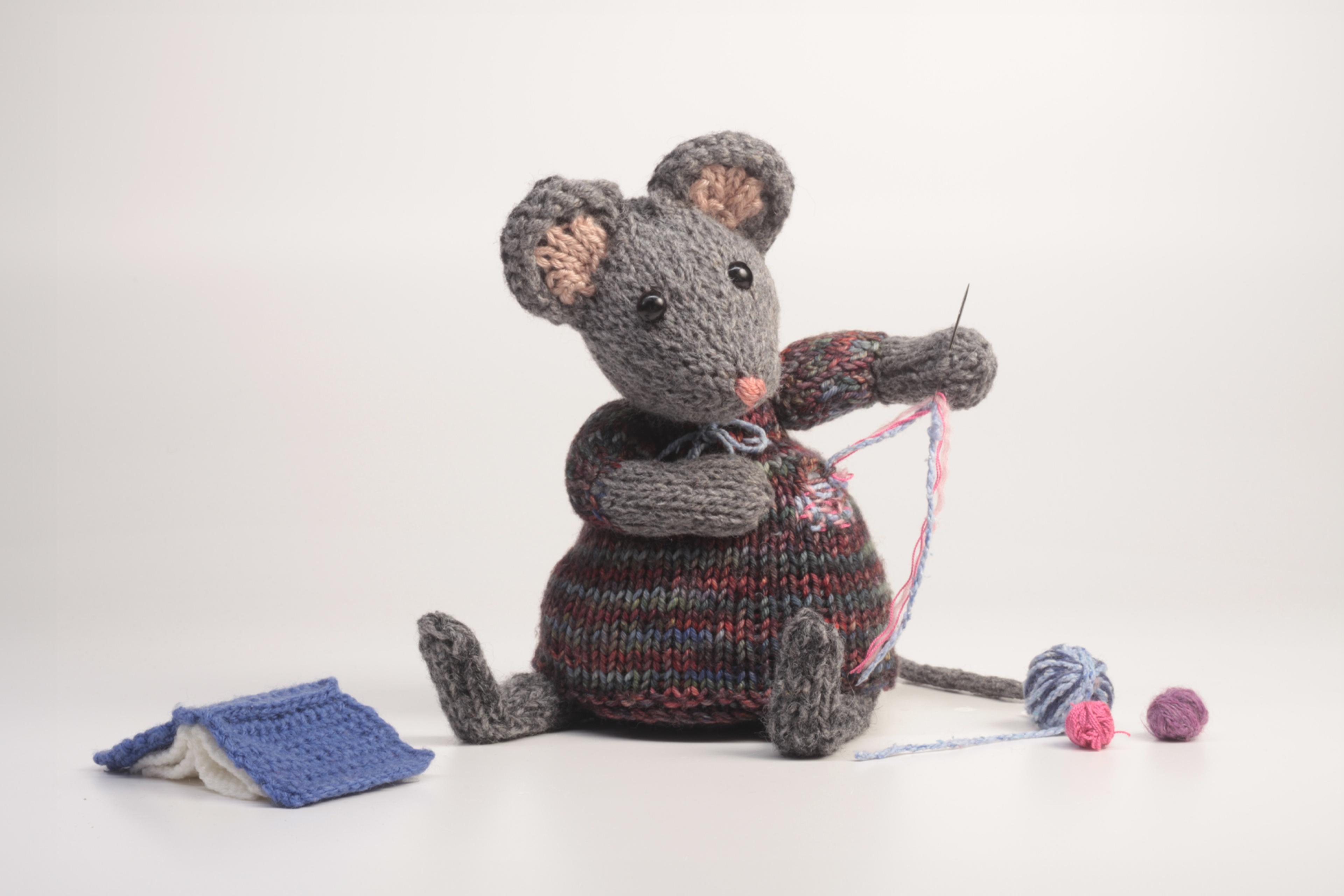 A knitted grey toy mouse holds a needle and repairs its own dress. Three balls of yarn lie nearby, alongside a small blue open book.
