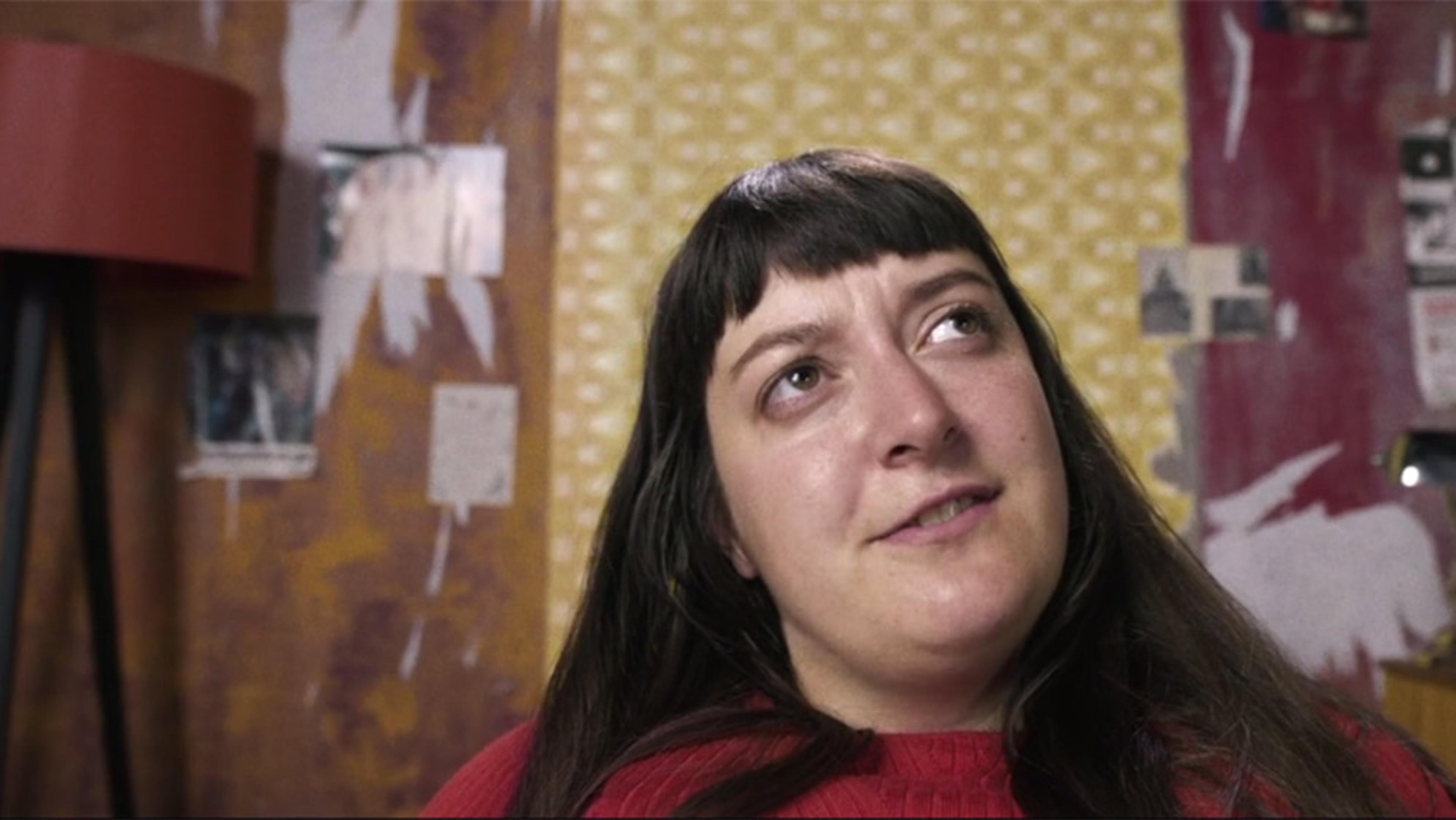 A woman with long dark hair and a fringe, wearing a red jumper, with a patterned yellow and red wall in the background.