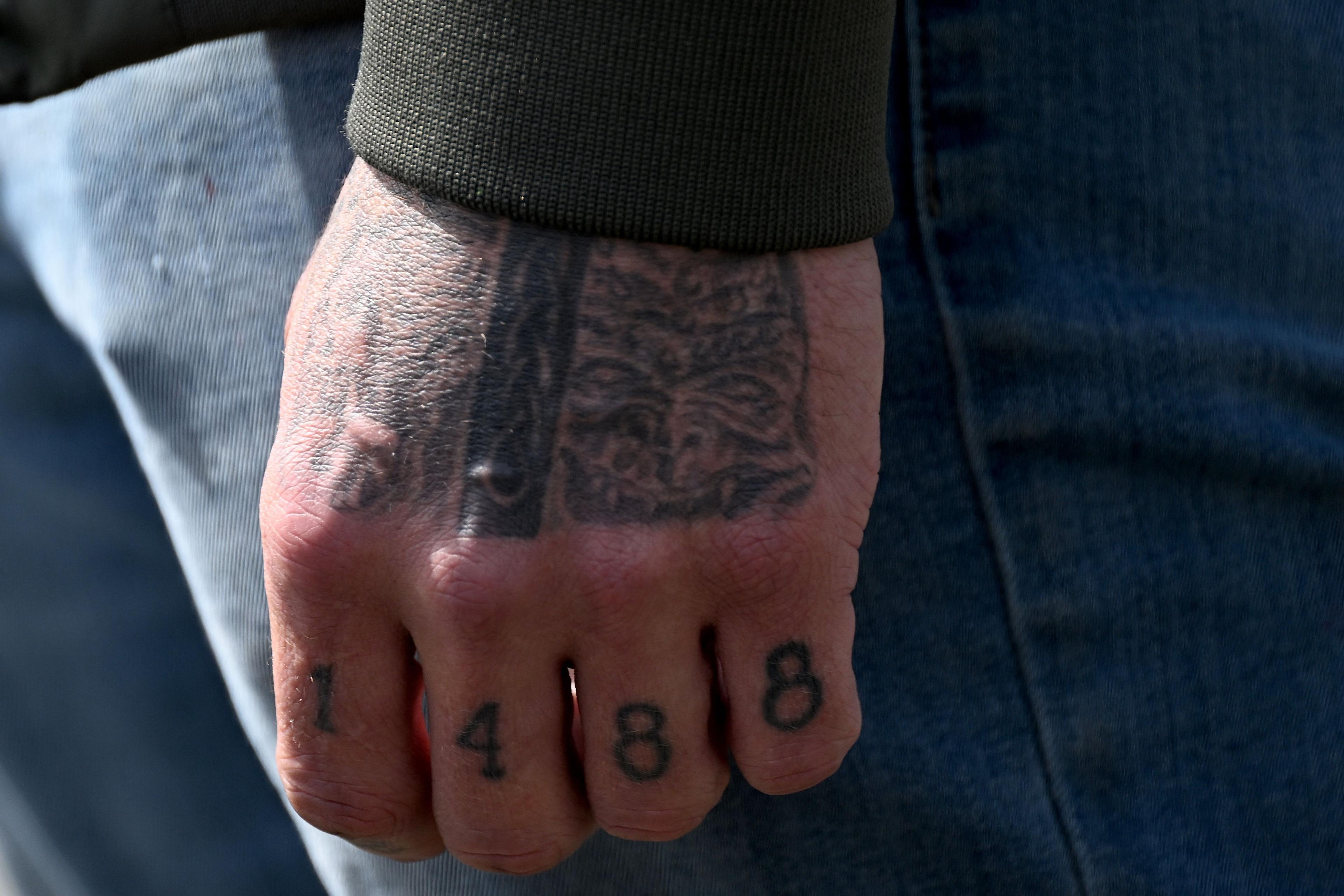 Photo of a hand with tattoos, including “1488” on the fingers, wearing a green sleeve and blue jeans in the background.