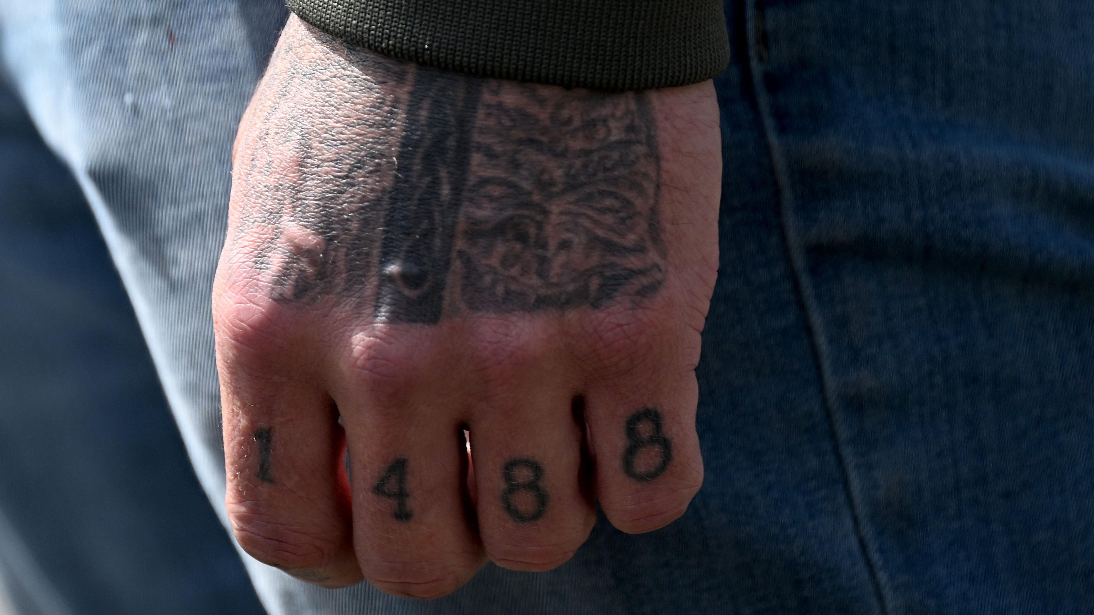 Photo of a hand with tattoos, including “1488” on the fingers, wearing a green sleeve and blue jeans in the background.