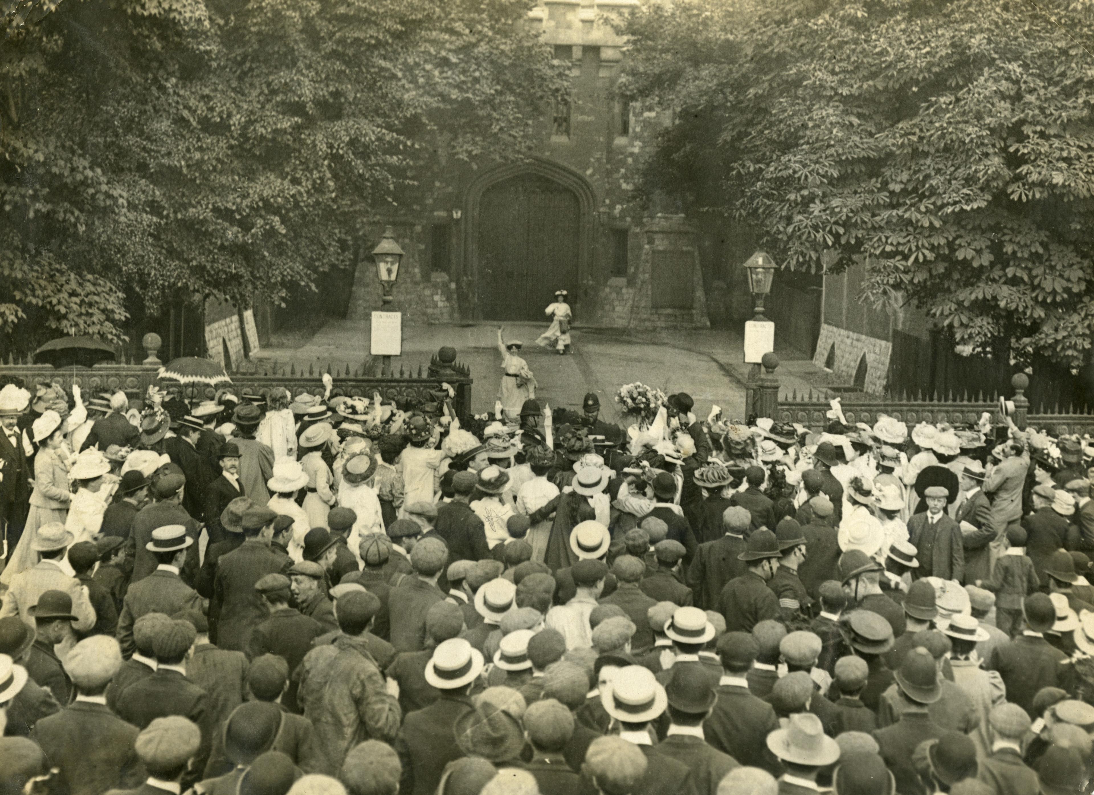 Historic photo of a large crowd gathered outside a gated building surrounded by trees, while two women in long dresses approach them from the building.