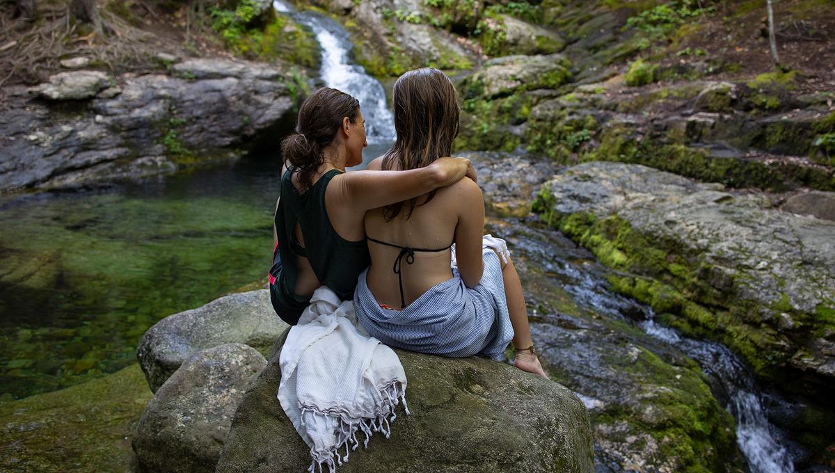 Two women wearing bathing suits sit on a rock by a waterfall in nature; one has her arm around the other. Towels are draped over the rock.