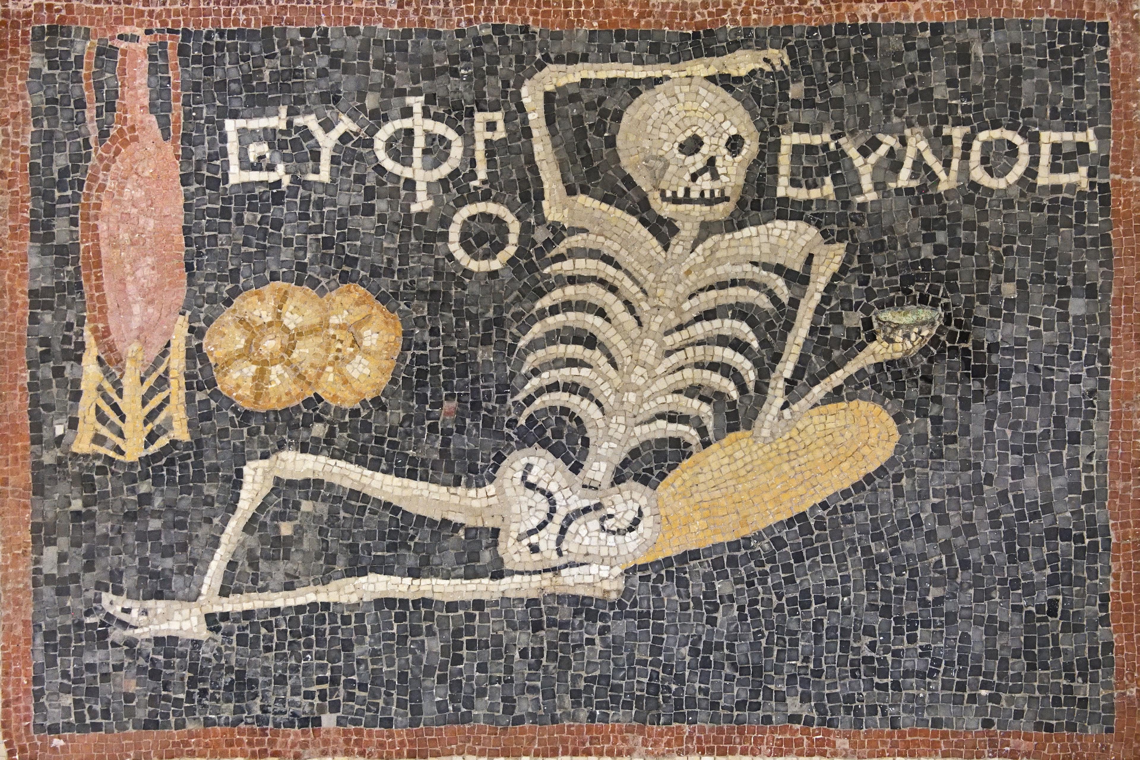 Ancient mosaic depicting a lounging skeleton with Greek text, an amphora, a loaf of bread and a platter on a dark background.