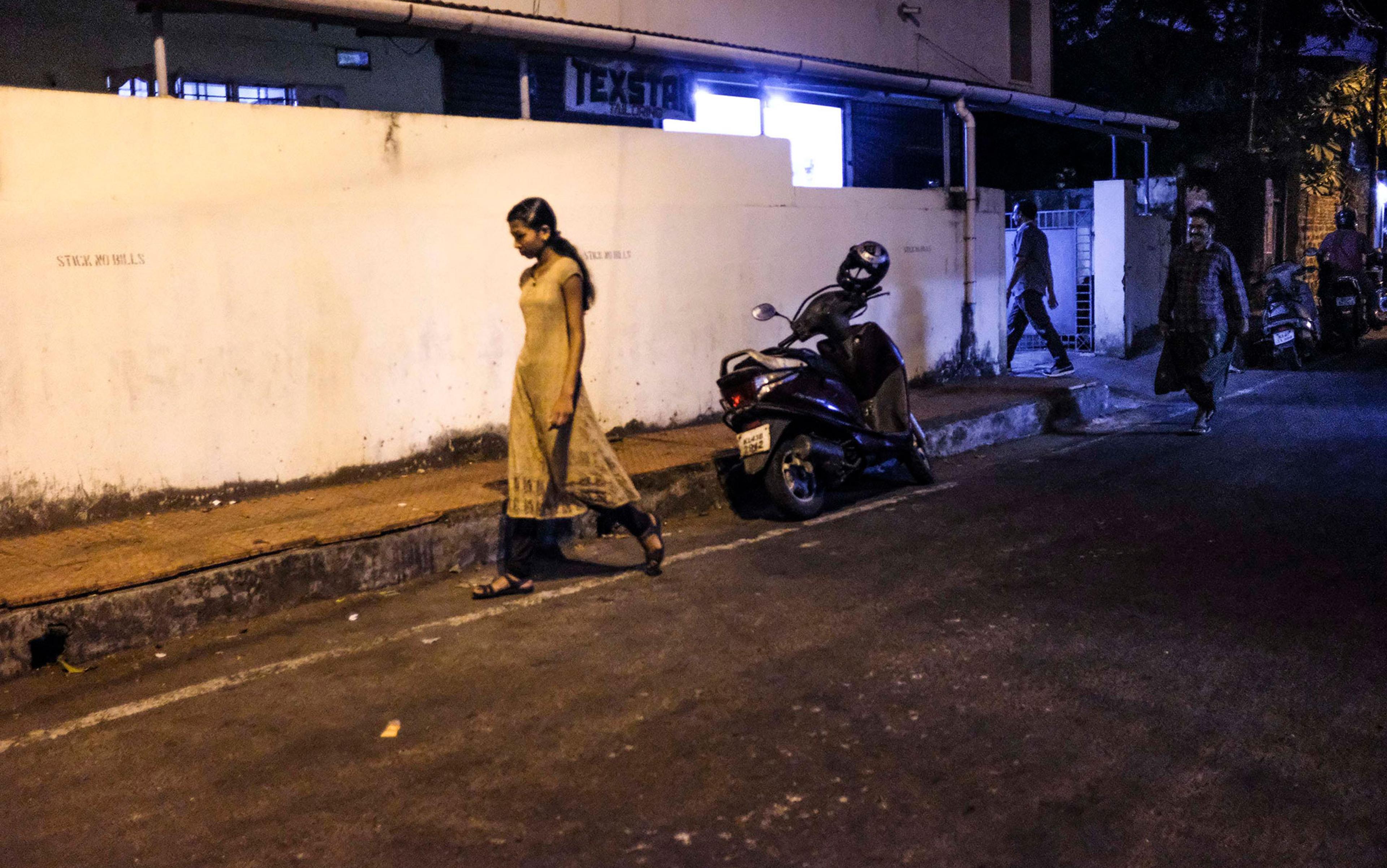 A woman walking on a dimly lit street pavement at night with a parked scooter and two men walking in the background, one appearing to follow her; the woman looks downcast, the man appears to be smiling.