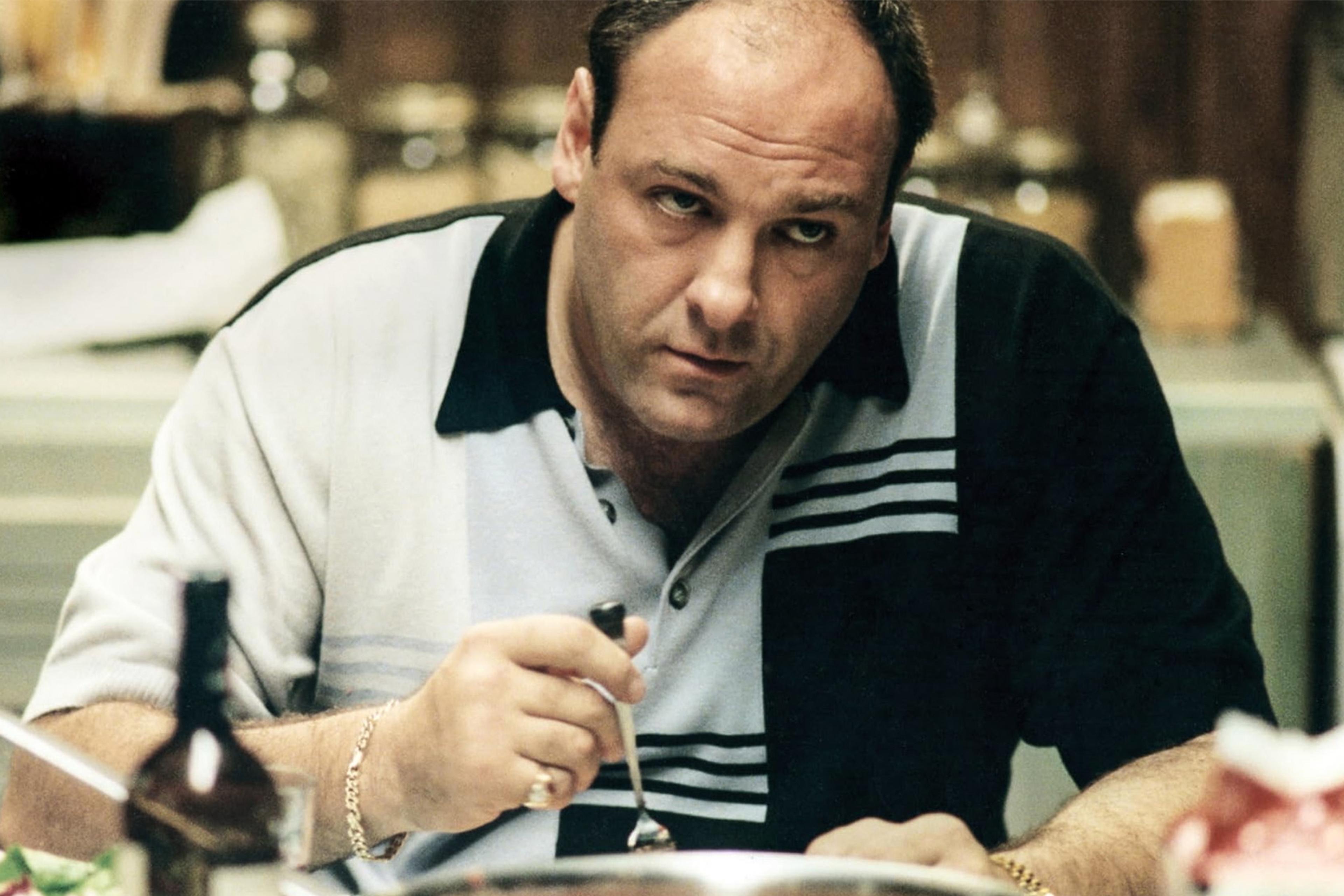 Photo of a man with a serious expression sitting at a table holding a fork in a kitchen setting.