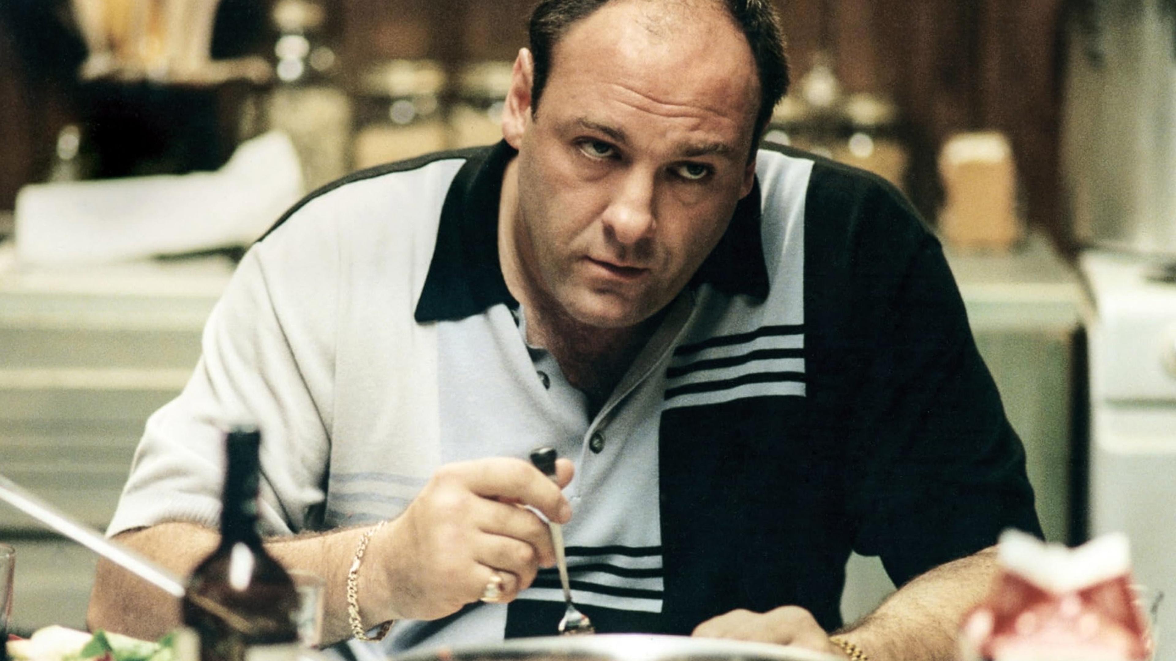 Photo of a man with a serious expression sitting at a table holding a fork in a kitchen setting.