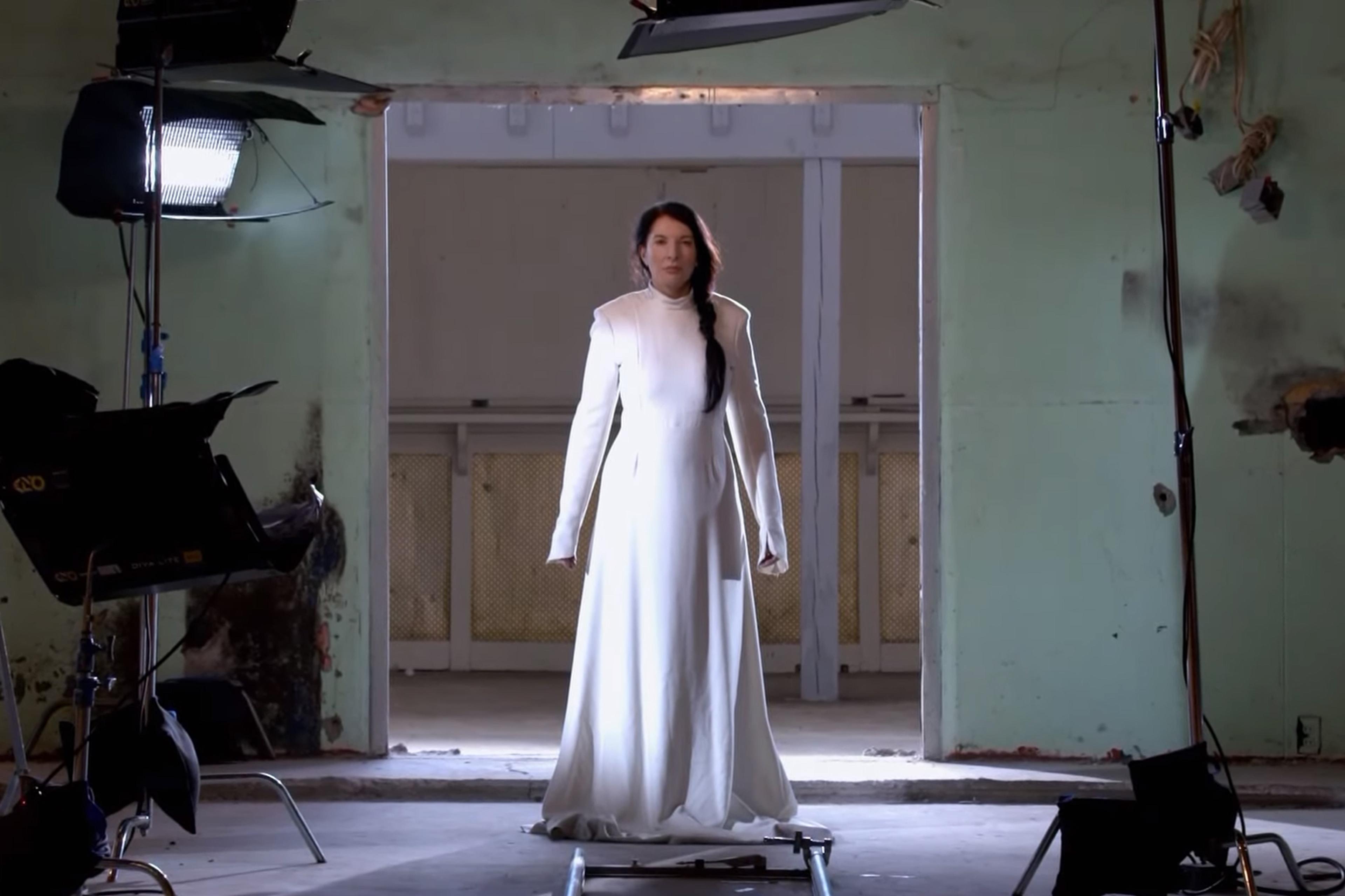The artist Marina Abramović in a long, white dress stands in an abandoned building, lit by studio lights. The walls are rundown and the floor has tracks for camera equipment. The woman faces forward, with her hair in a side braid, and the atmosphere is dramatic and moody.