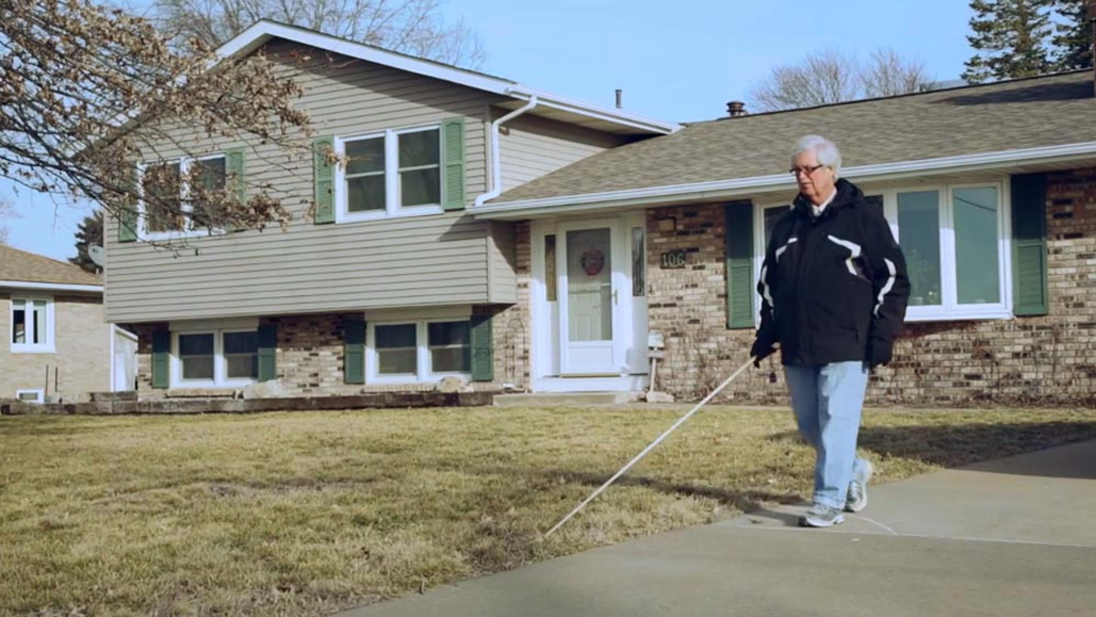 An older man with a walking stick, walking on a pavement in front of a house with green shutters on a sunny day.