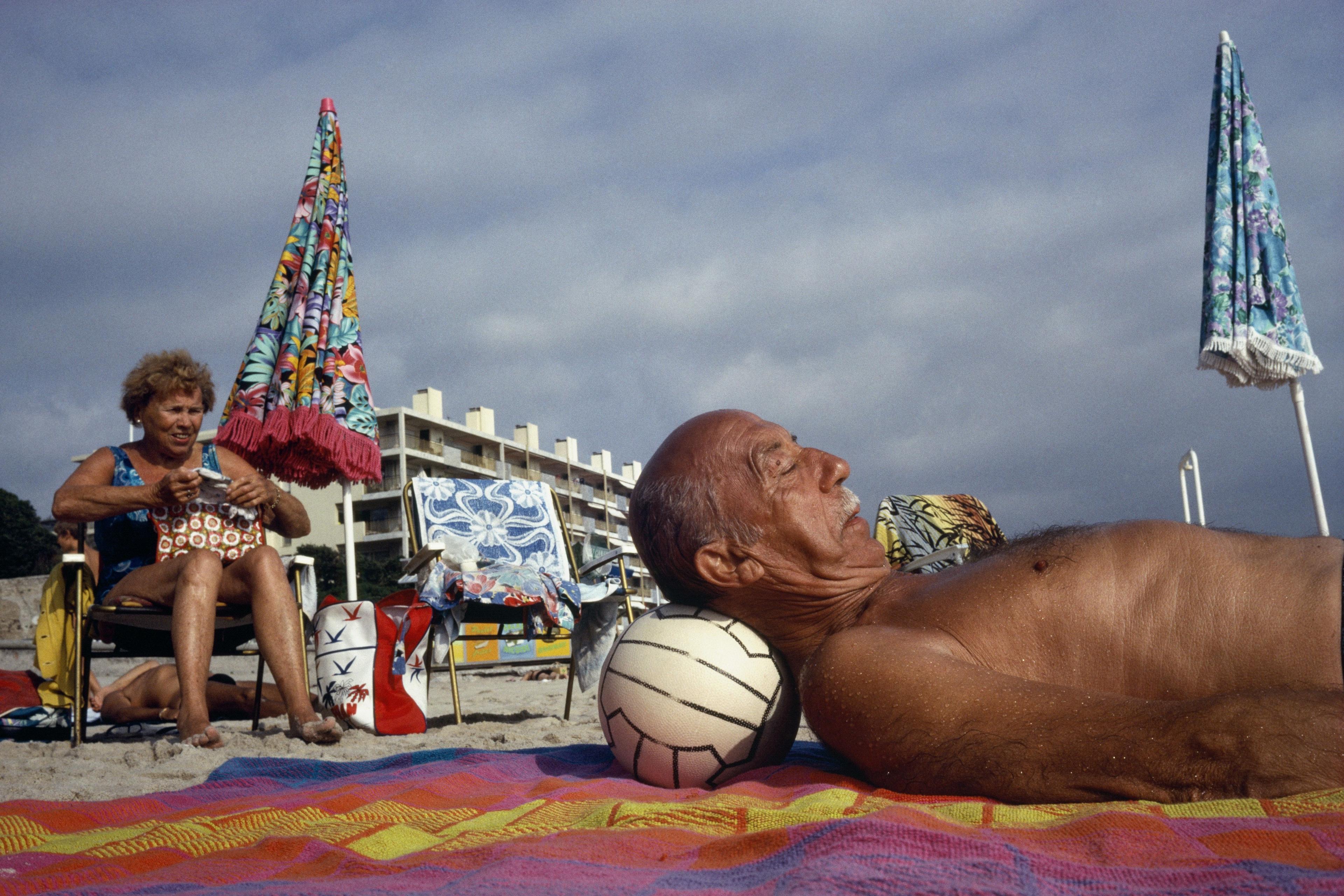 Photo of a man lounging on a beach towel with a ball as a pillow a woman sits nearby with colourful umbrellas and buildings.
