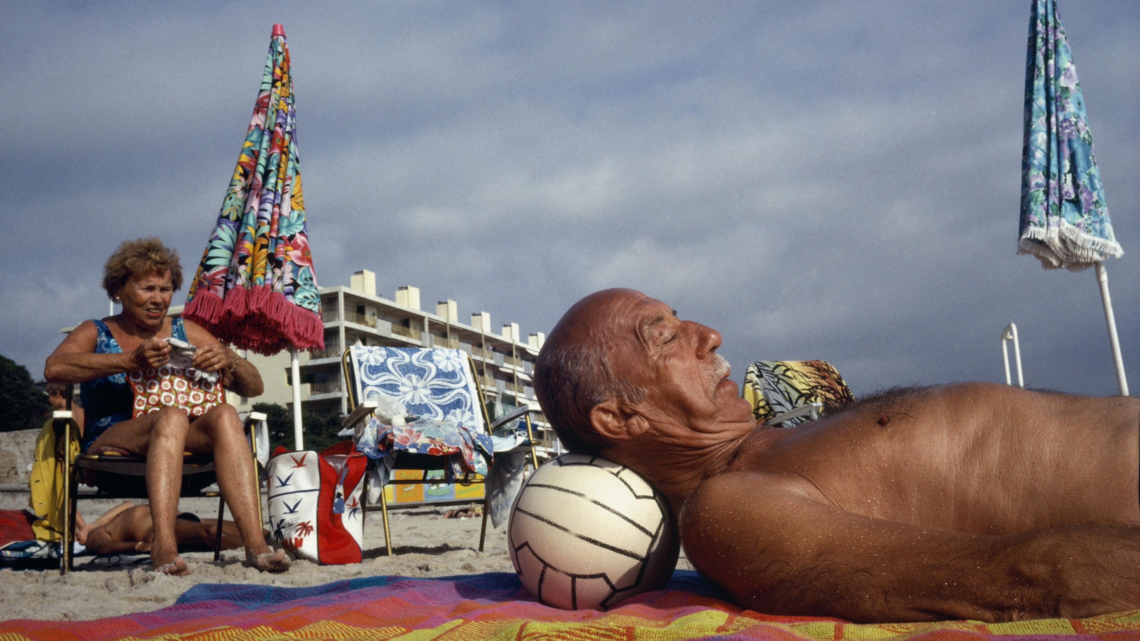 Photo of a man lounging on a beach towel with a ball as a pillow a woman sits nearby with colourful umbrellas and buildings.