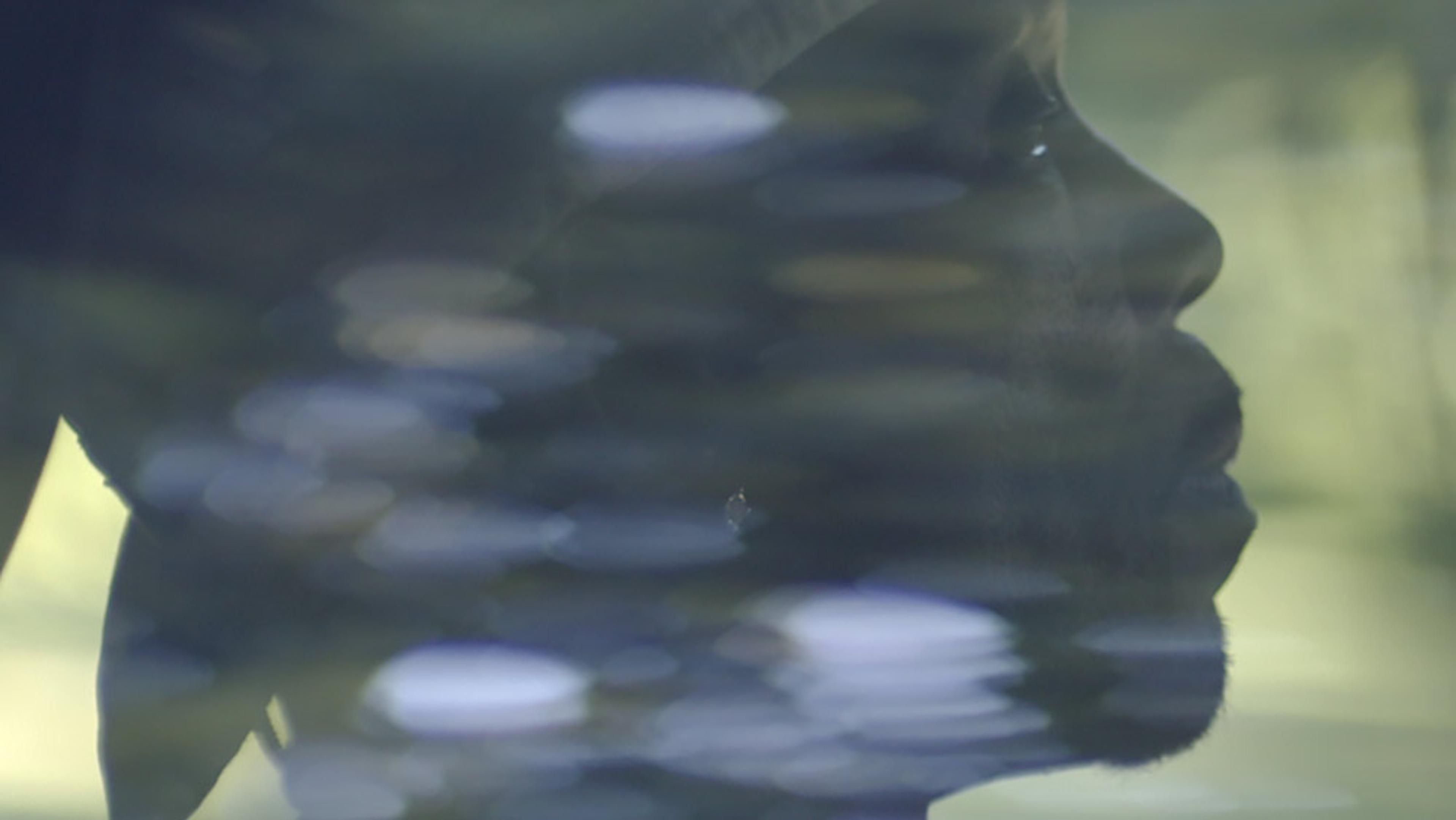 A man’s side profile viewed through a window with blurred reflections, capturing a pensive and serene atmosphere.