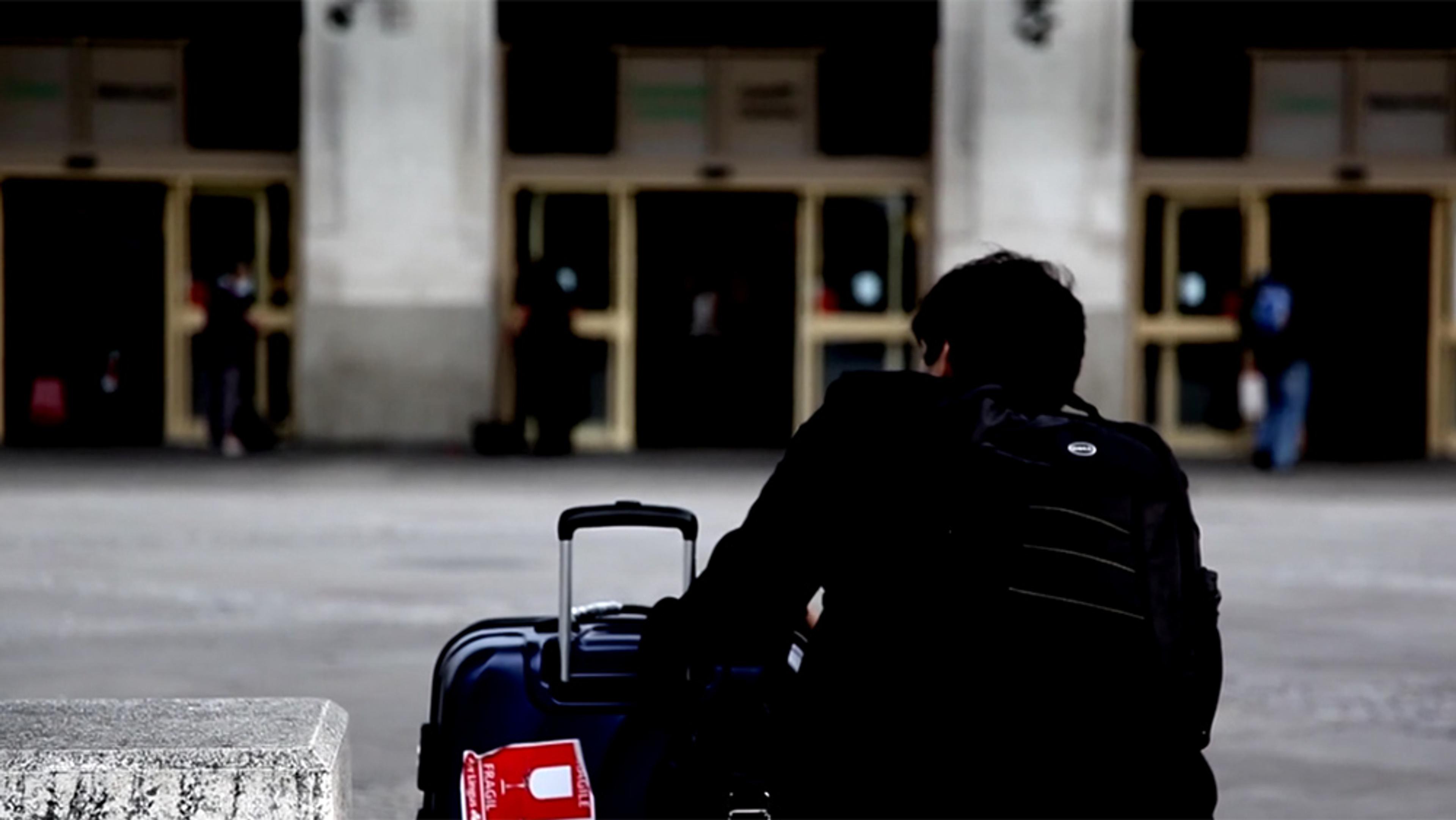 A person in dark clothing sitting with luggage outside a building entrance, with other figures in the background.