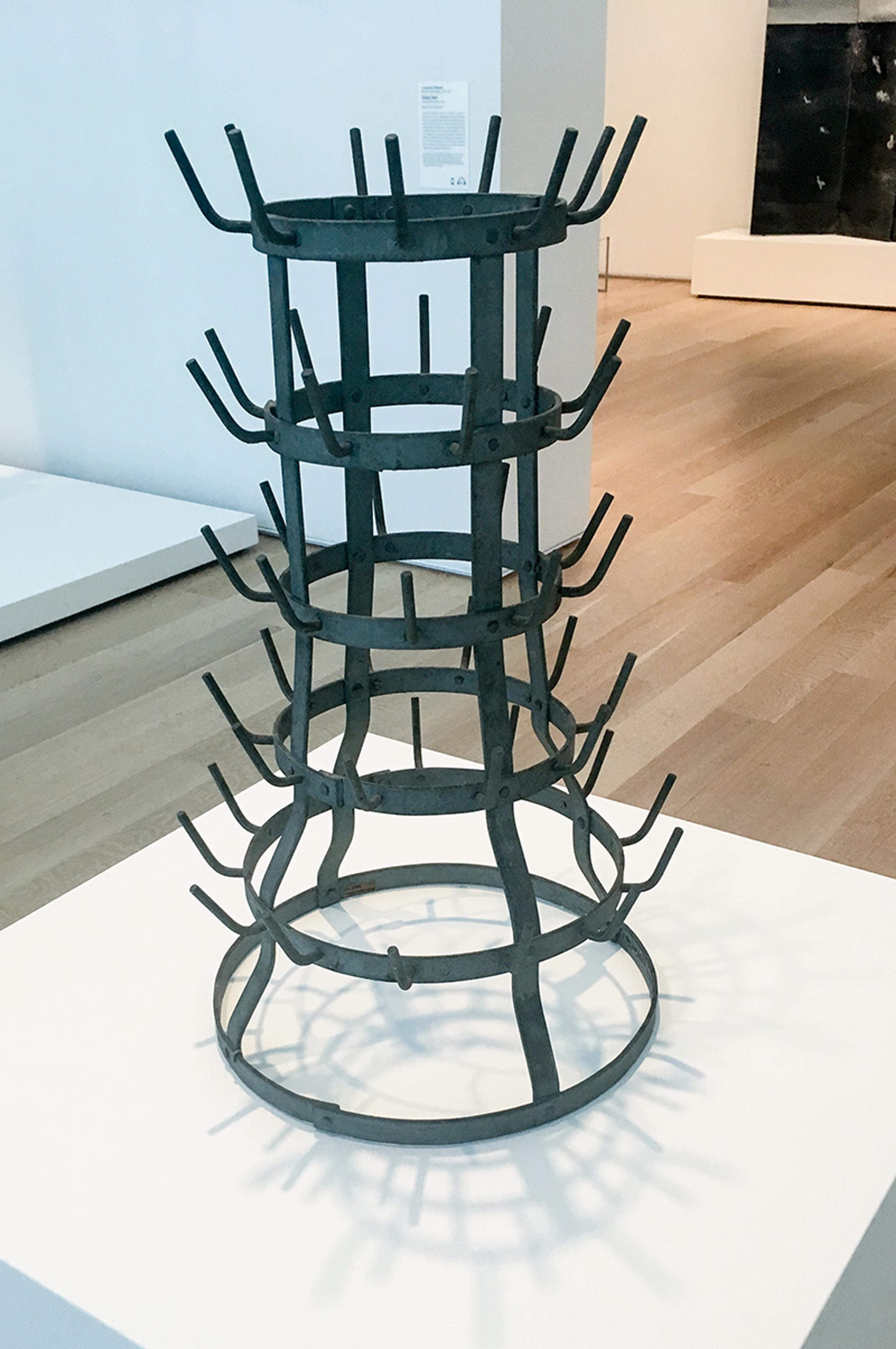 A metal bottle-drying rack with multiple levels and protruding pegs, displayed in an art gallery on a white pedestal.