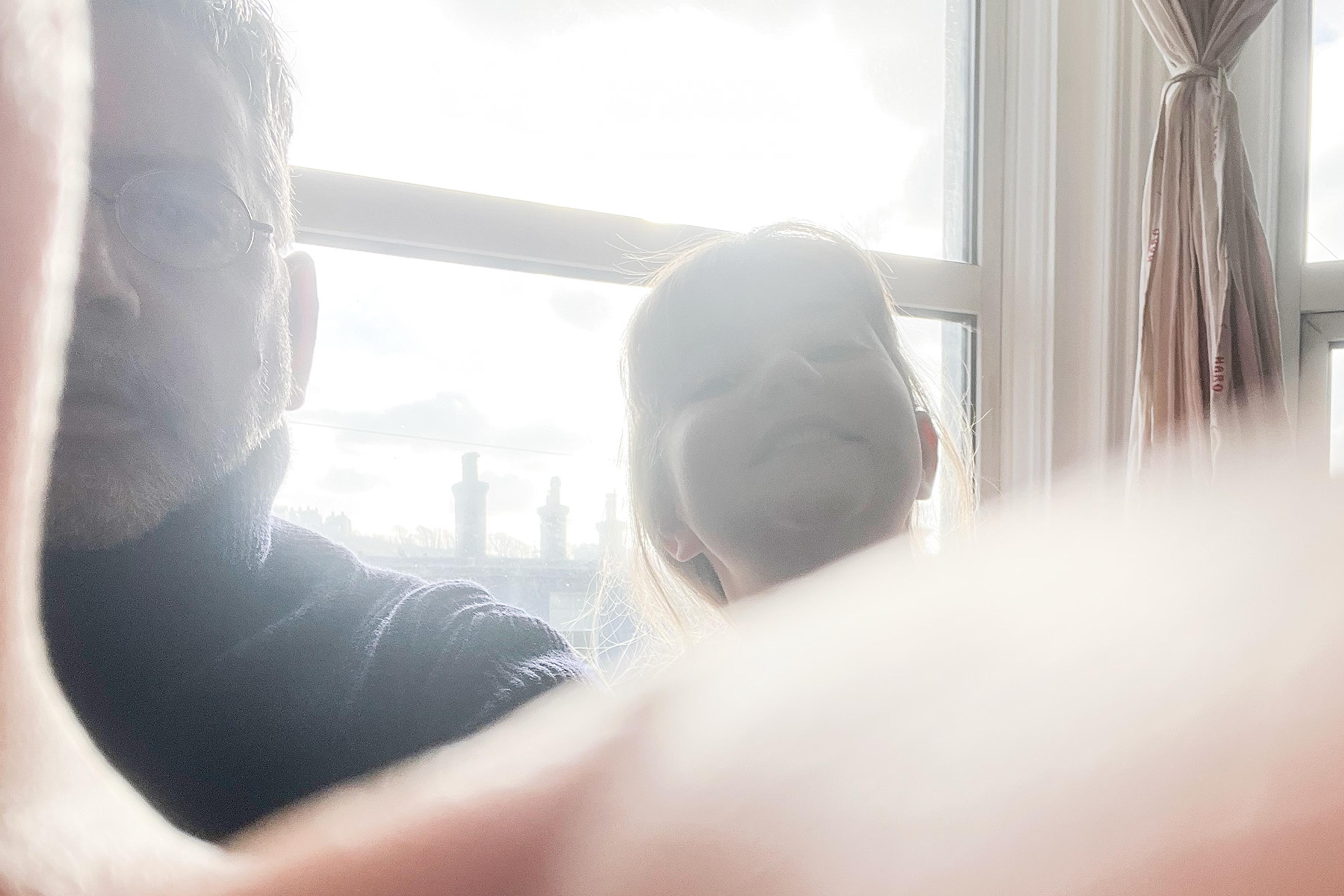 Photo of two people partially obscured, with sunlight streaming through a window behind them and a thumb in the foreground.