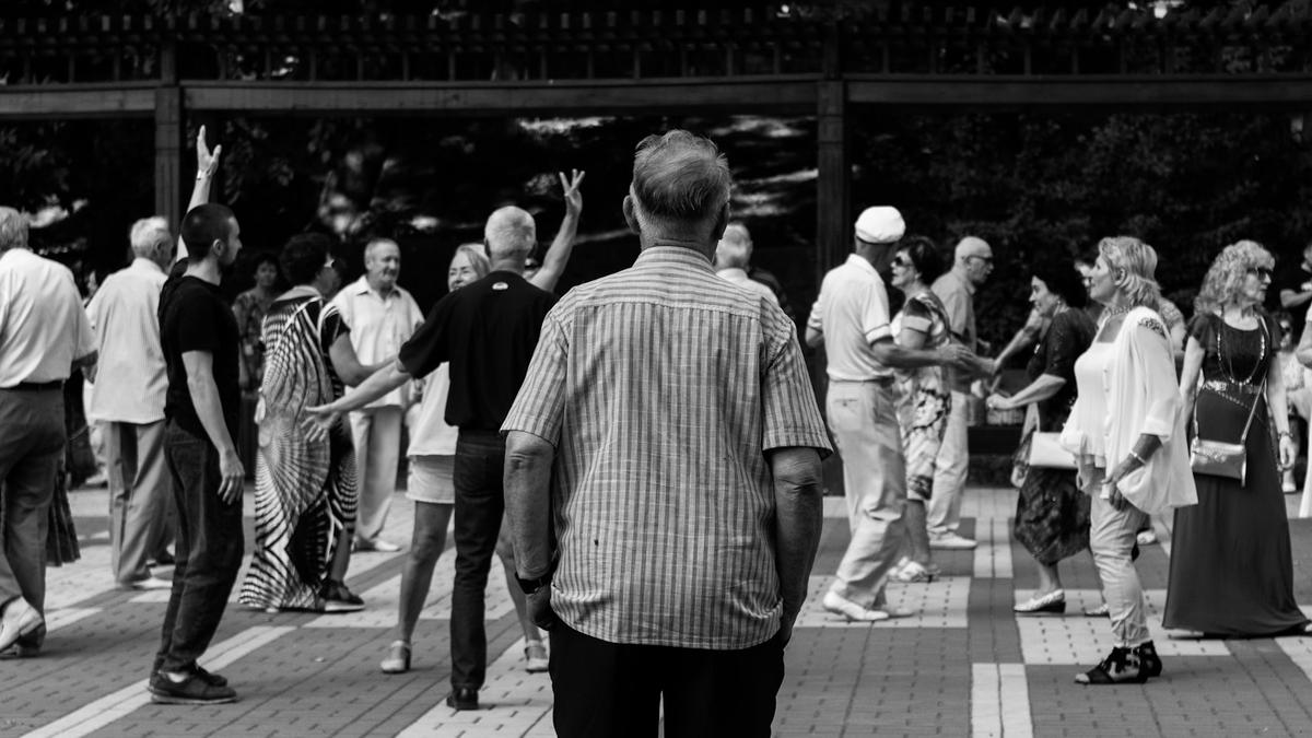 Black and white photo of a group of people dancing outdoors, with a man in a striped shirt in the foreground.