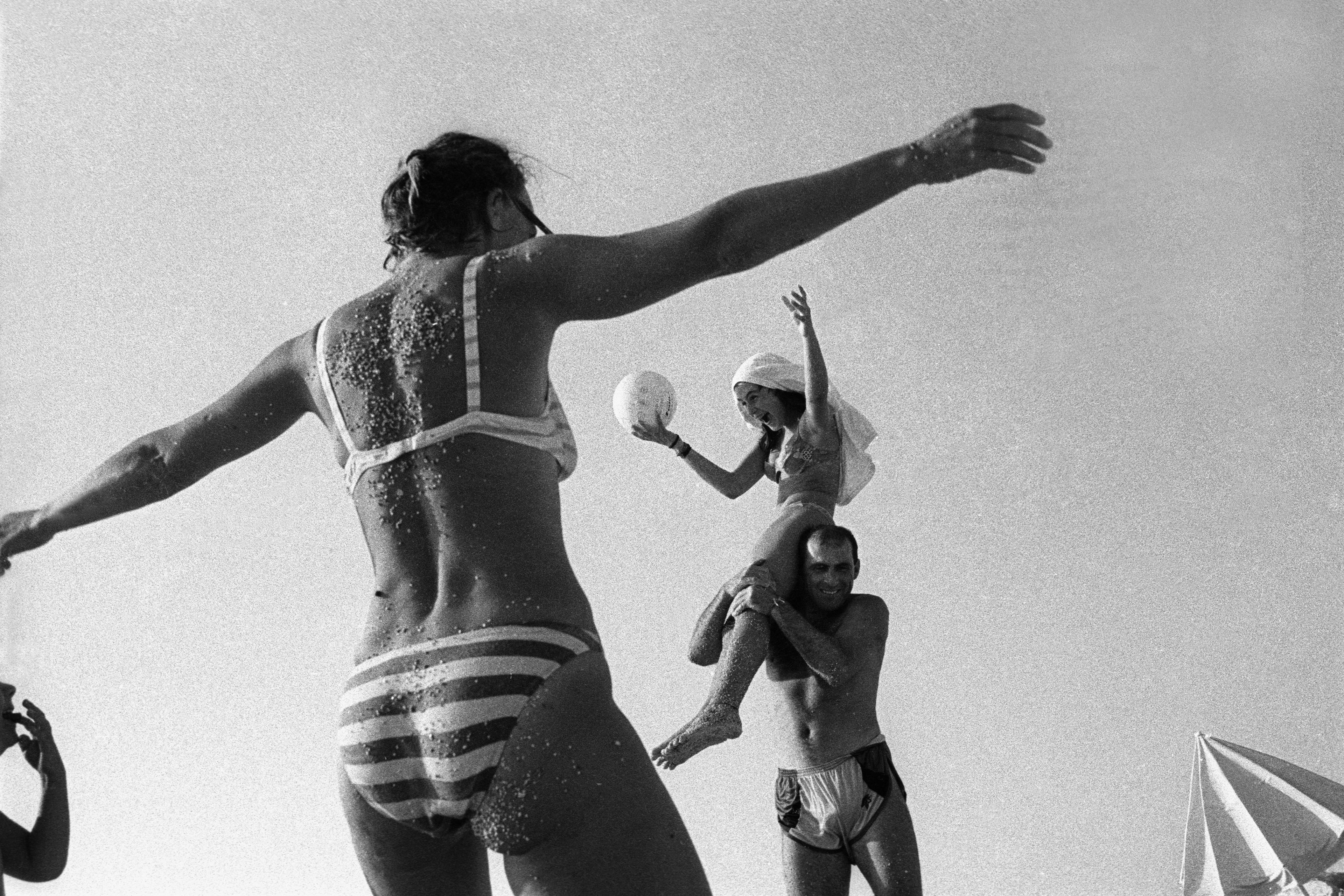 Black and white photo of people playing on a beach. A woman sits on a man’s shoulders holding a ball, others move around them.