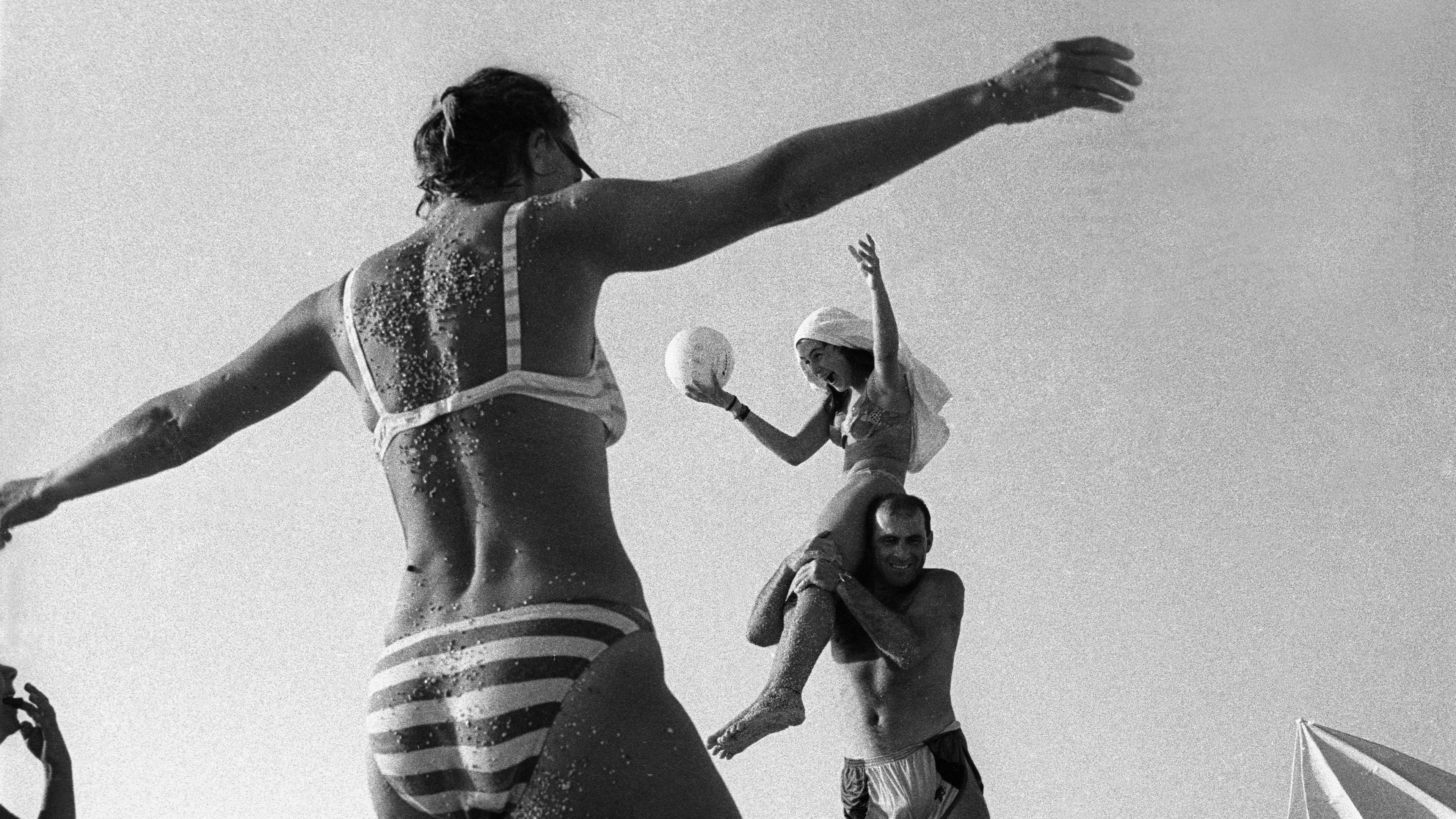 Black and white photo of people playing on a beach. A woman sits on a man’s shoulders holding a ball, others move around them.