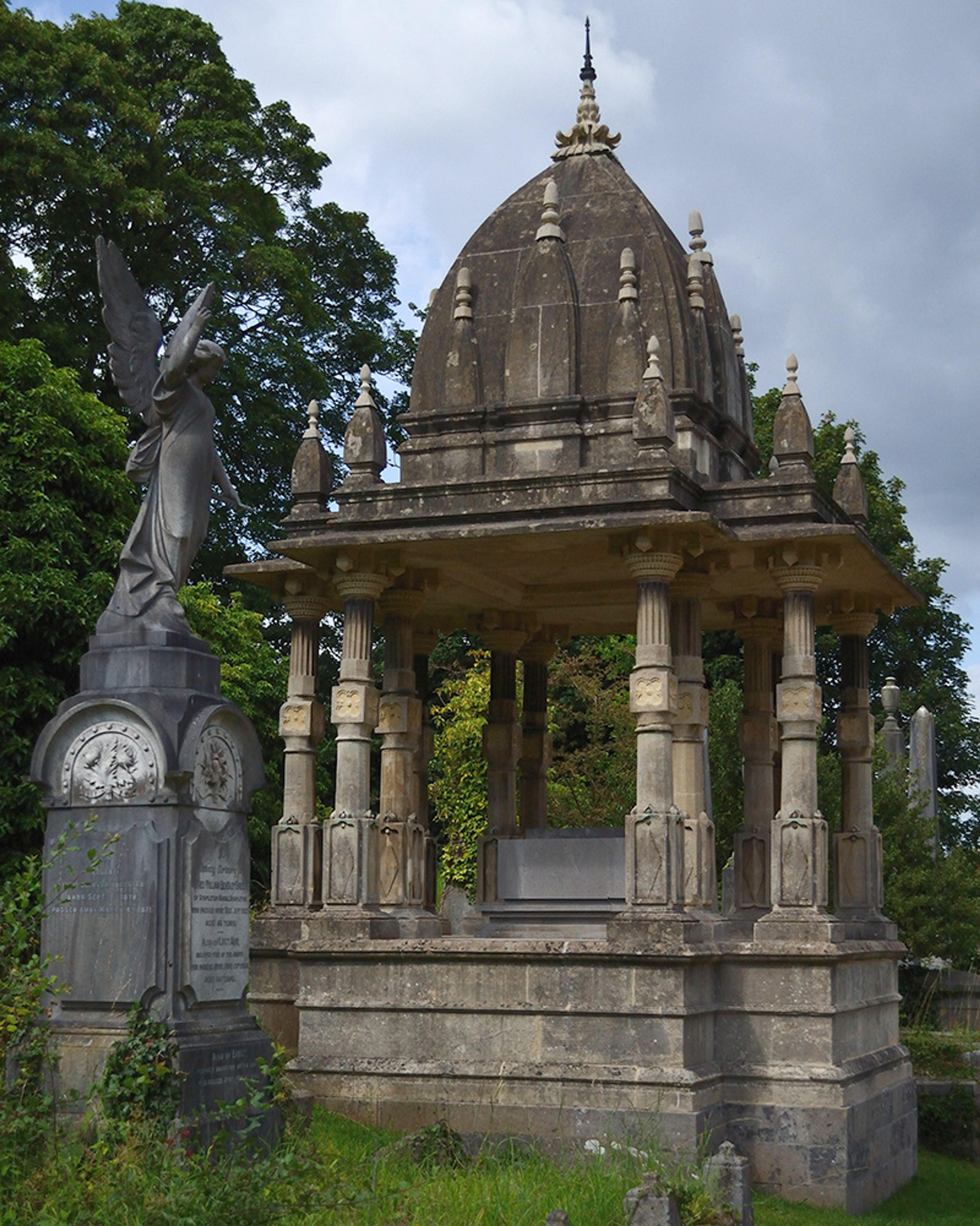 An ornate stone tomb with columns and a domed roof, accompanied by an angel statue, set in a cemetery.