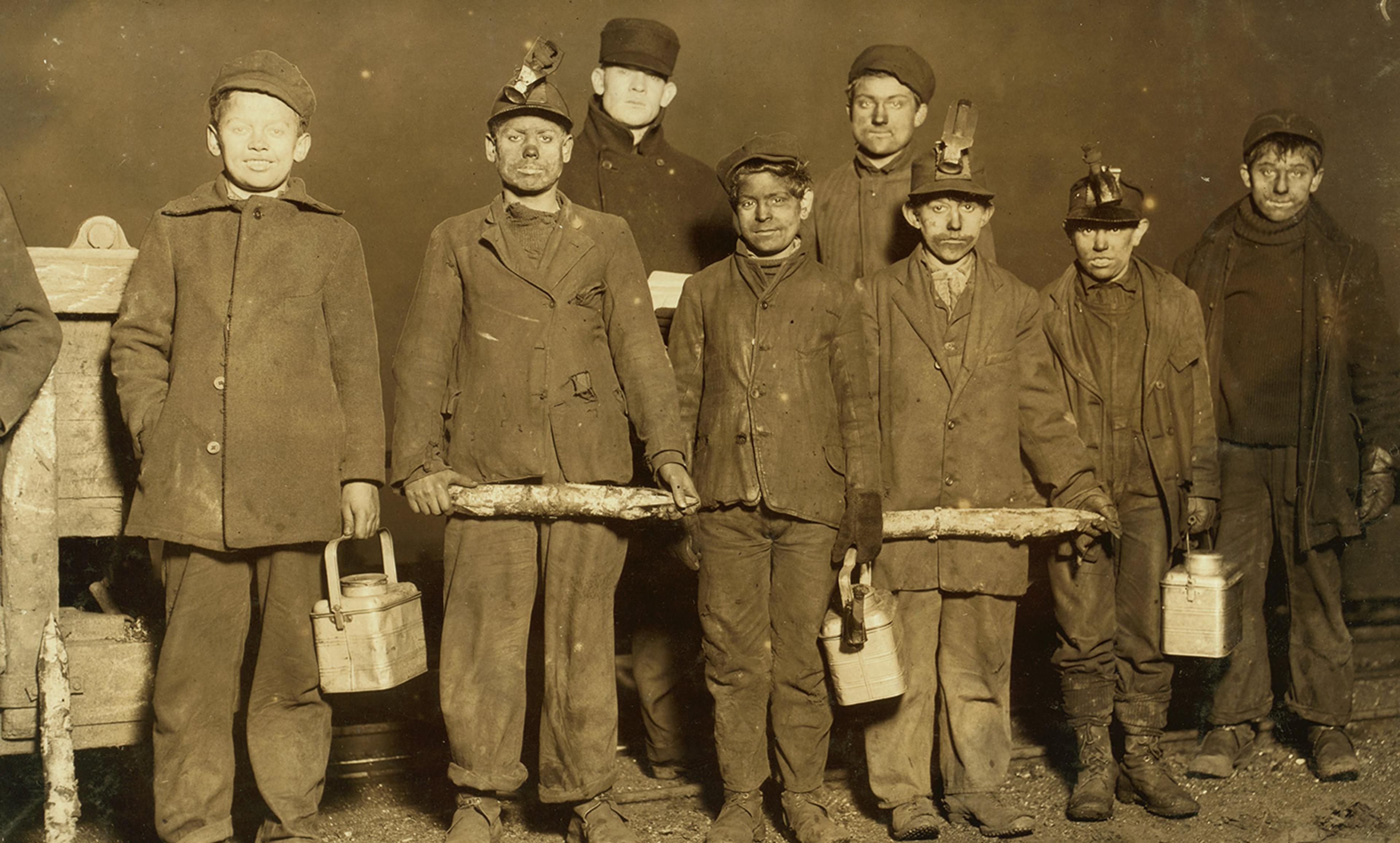 A vintage photograph of a group of young coal miners in work clothes holding lunch pails, taken in the early 20th century.