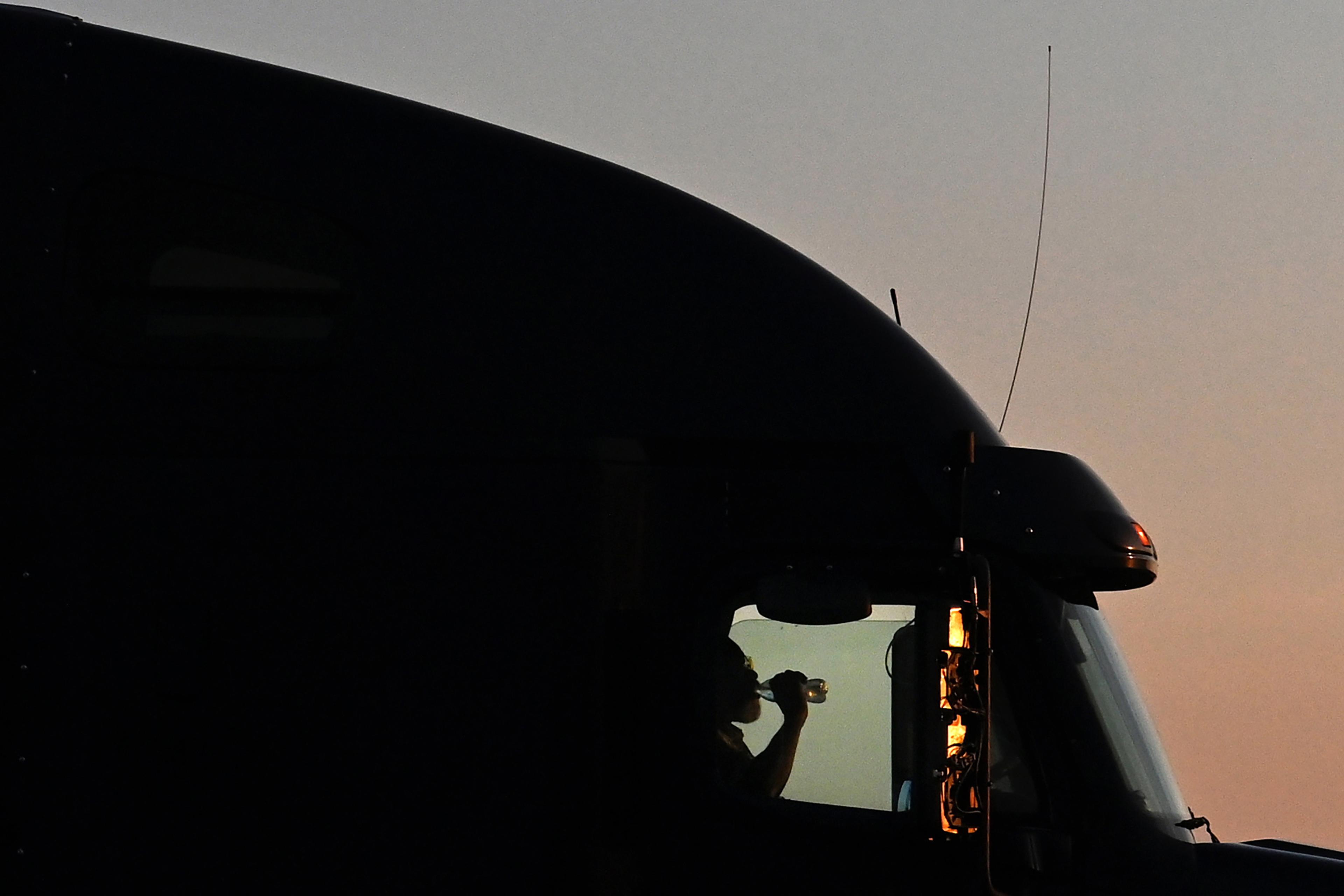 Silhouette photo of a person drinking from a bottle inside a lorry cab against a sunset sky.
