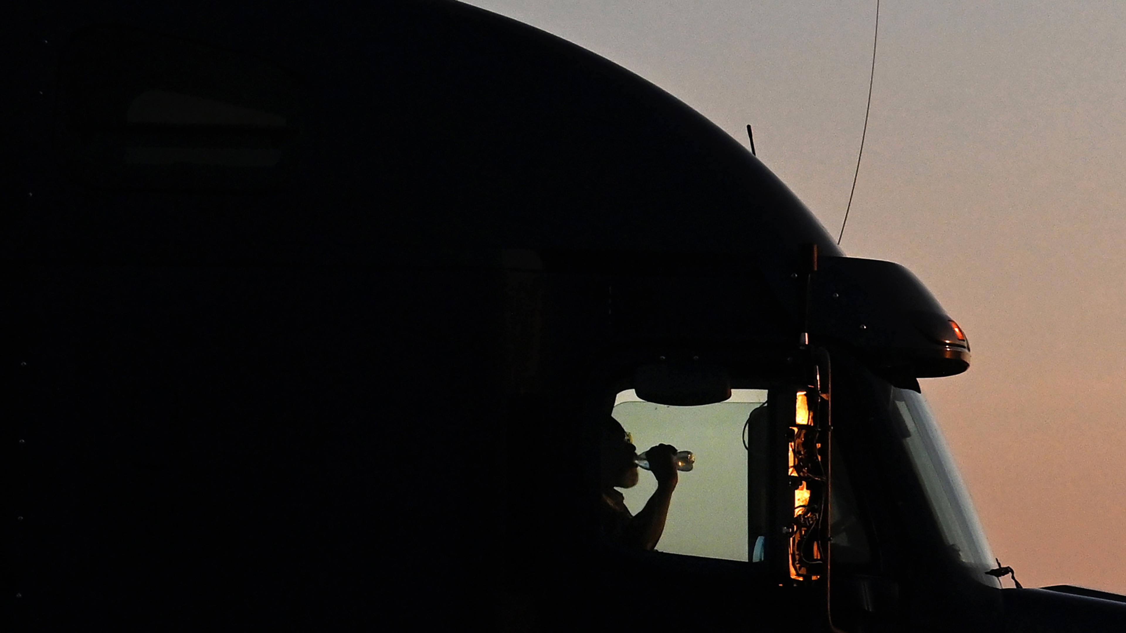 Silhouette photo of a person drinking from a bottle inside a lorry cab against a sunset sky.
