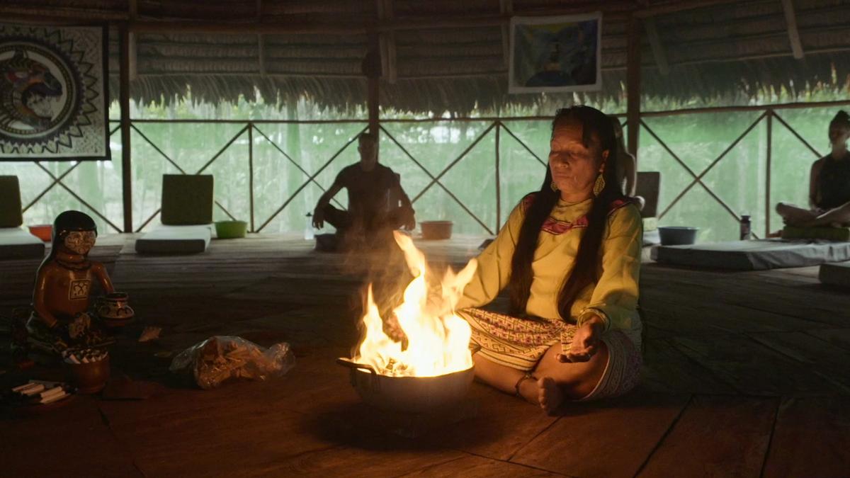 Photo of a woman meditating by a fire in a dimly lit room with artwork on the walls and others seated in the background.