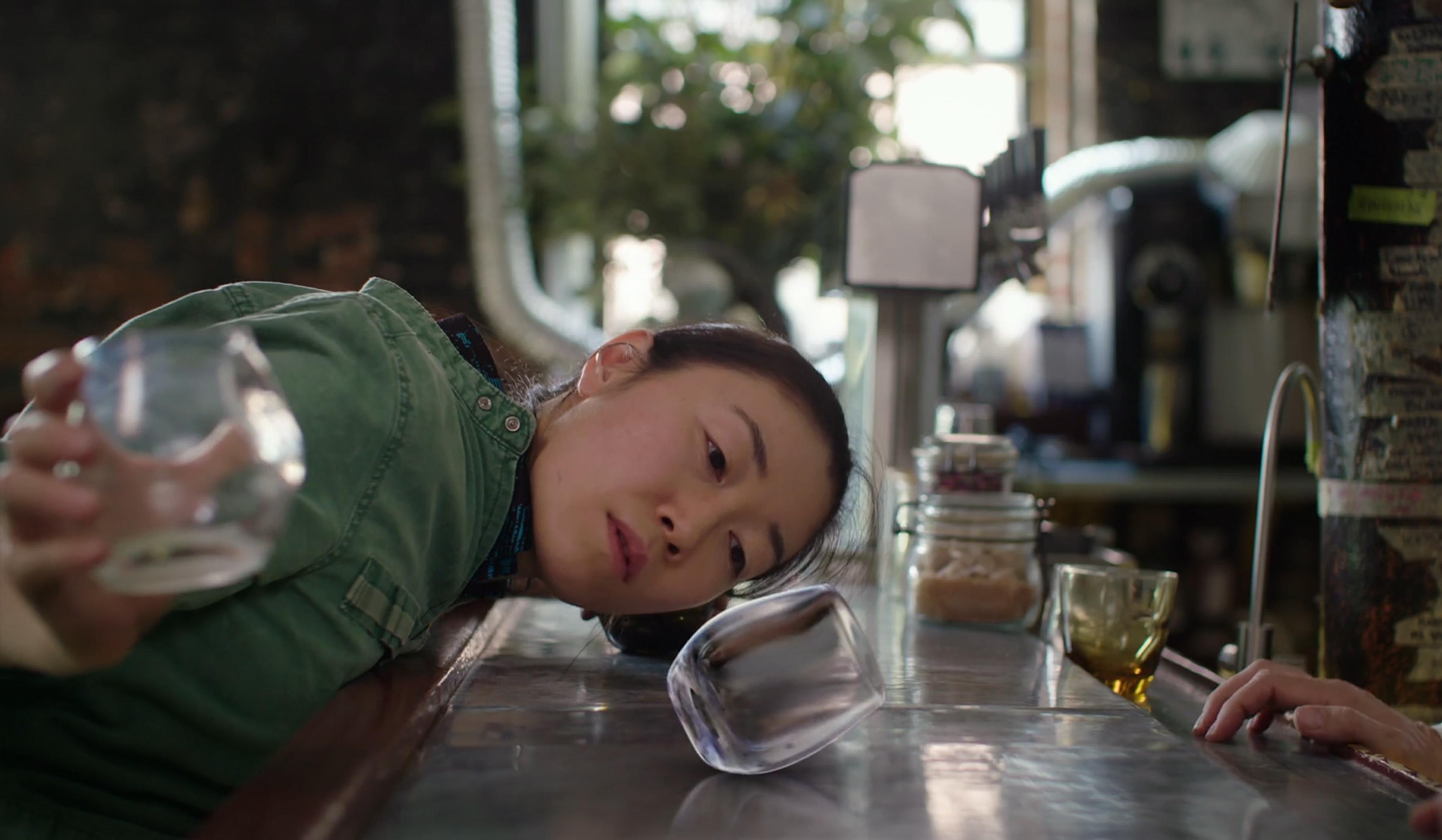 A woman leaning on a bar counter holding a glass and watching another glass tumble. Background shows a cafe interior.