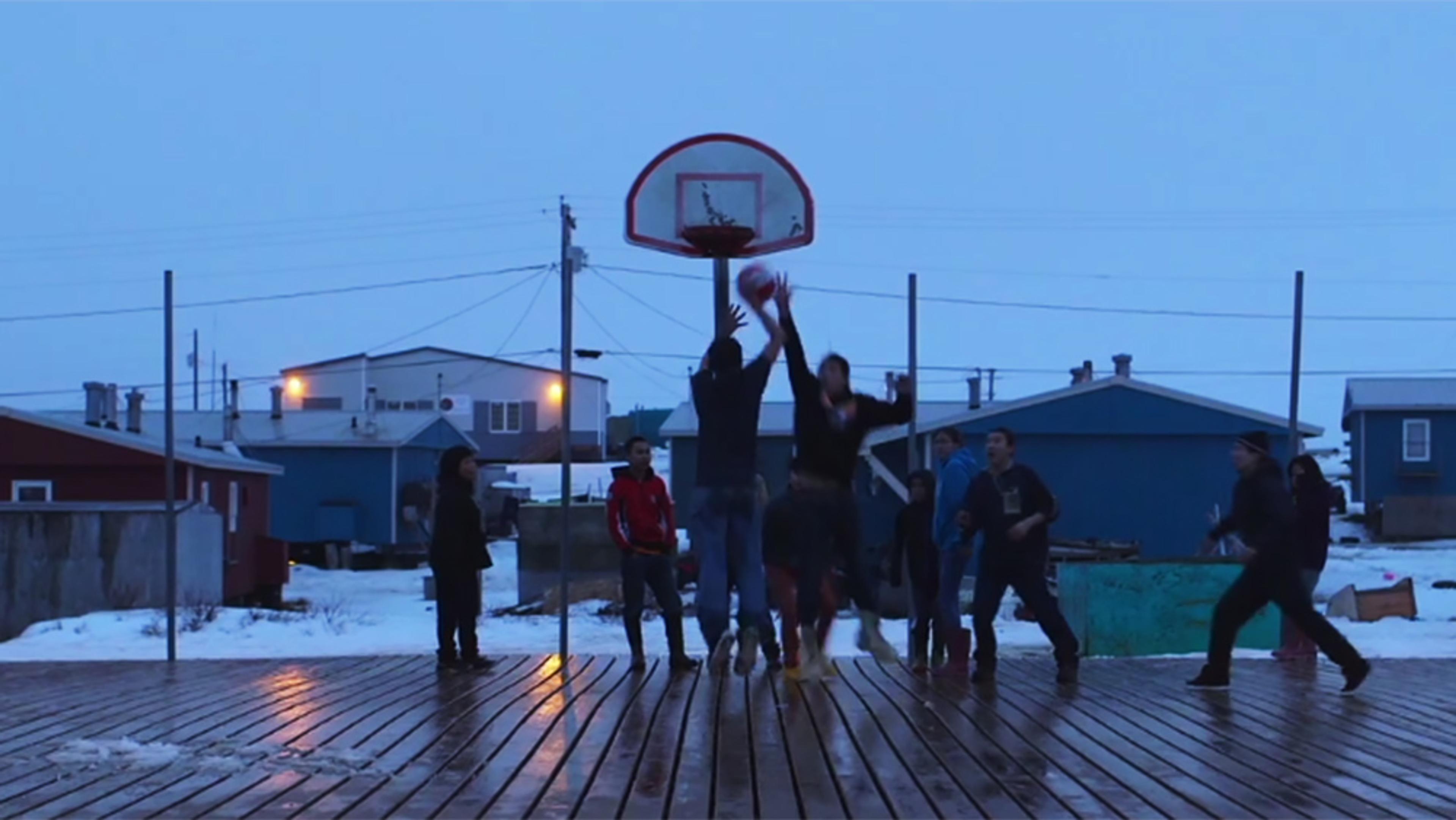 A group of people playing basketball outdoors on a wooden court with snow around; houses and power lines are in the background.