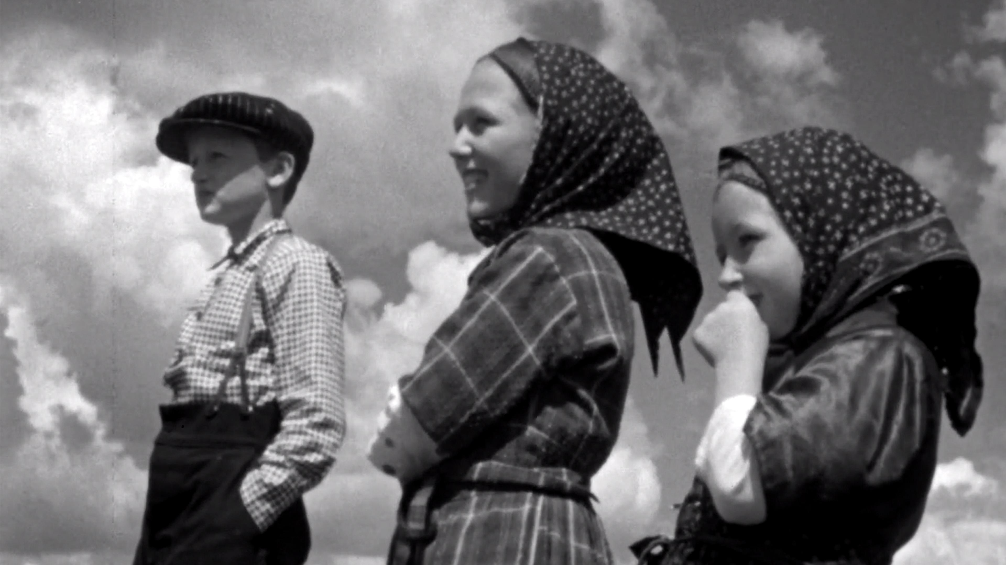 Black and white photo of three children standing outdoors under a cloudy sky. The boy on the left wears a flat cap and checked shirt with trousers, while the two girls on the right wear patterned headscarves and dresses, smiling and looking into the distance.
