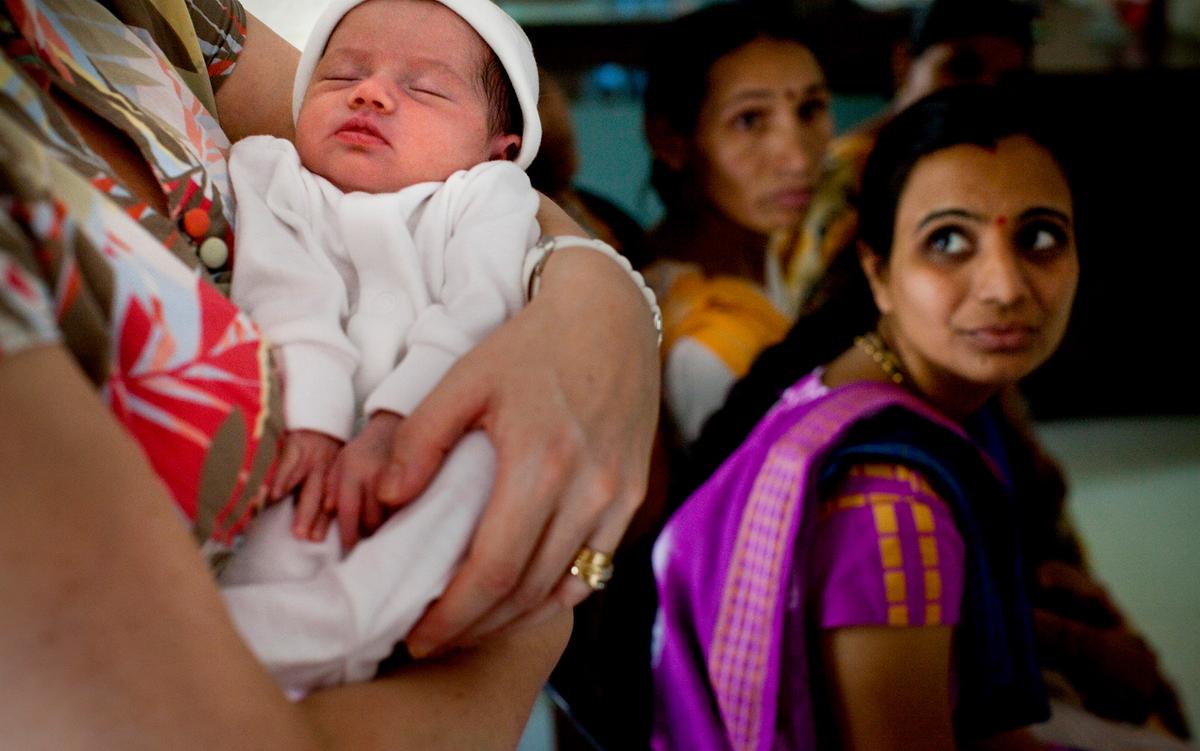 In the foreground a European mother cradles a newborn baby as an Indian woman looks on from the right