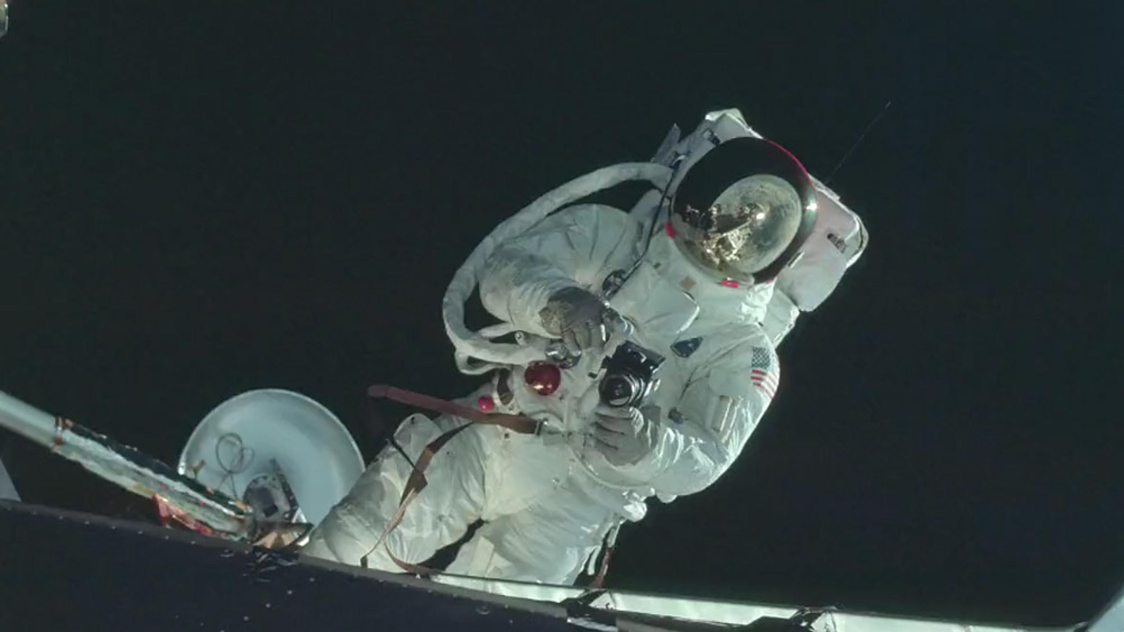 An astronaut in a spacesuit during a spacewalk holding a camera with Earth visible in the reflection of the helmet visor.