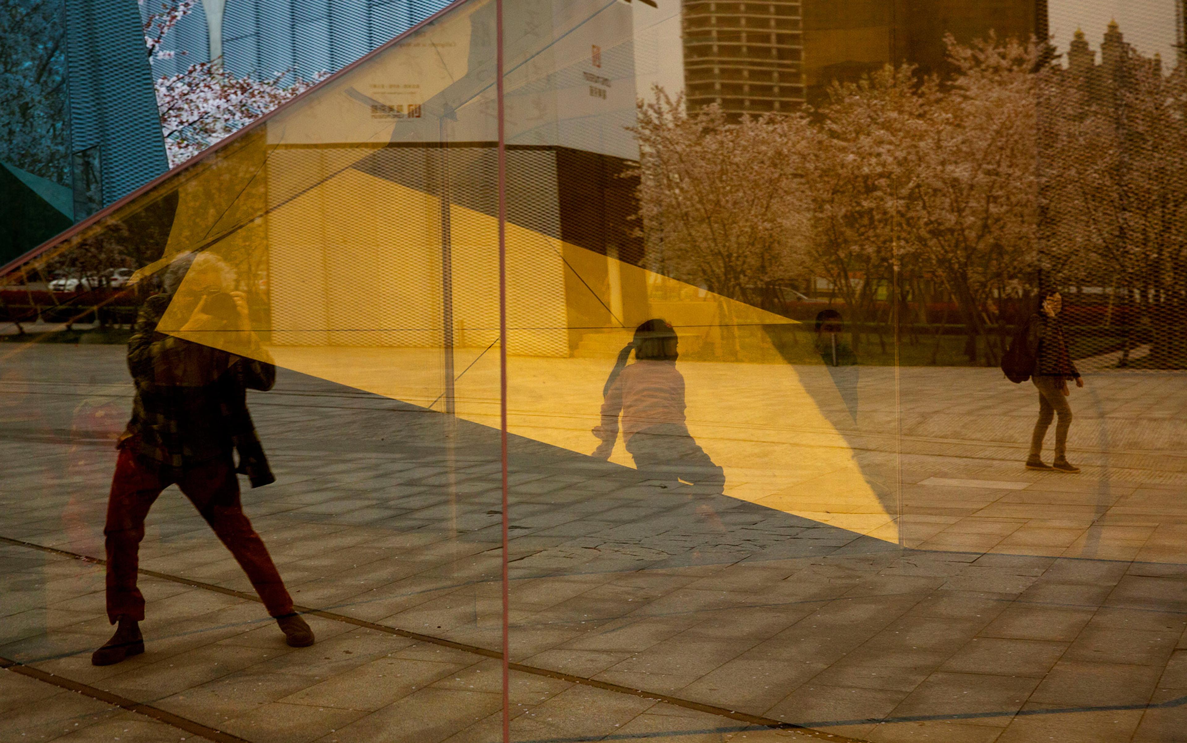 Photo showing reflections of people, trees and colourful geometric shapes on a glass surface in an urban setting.