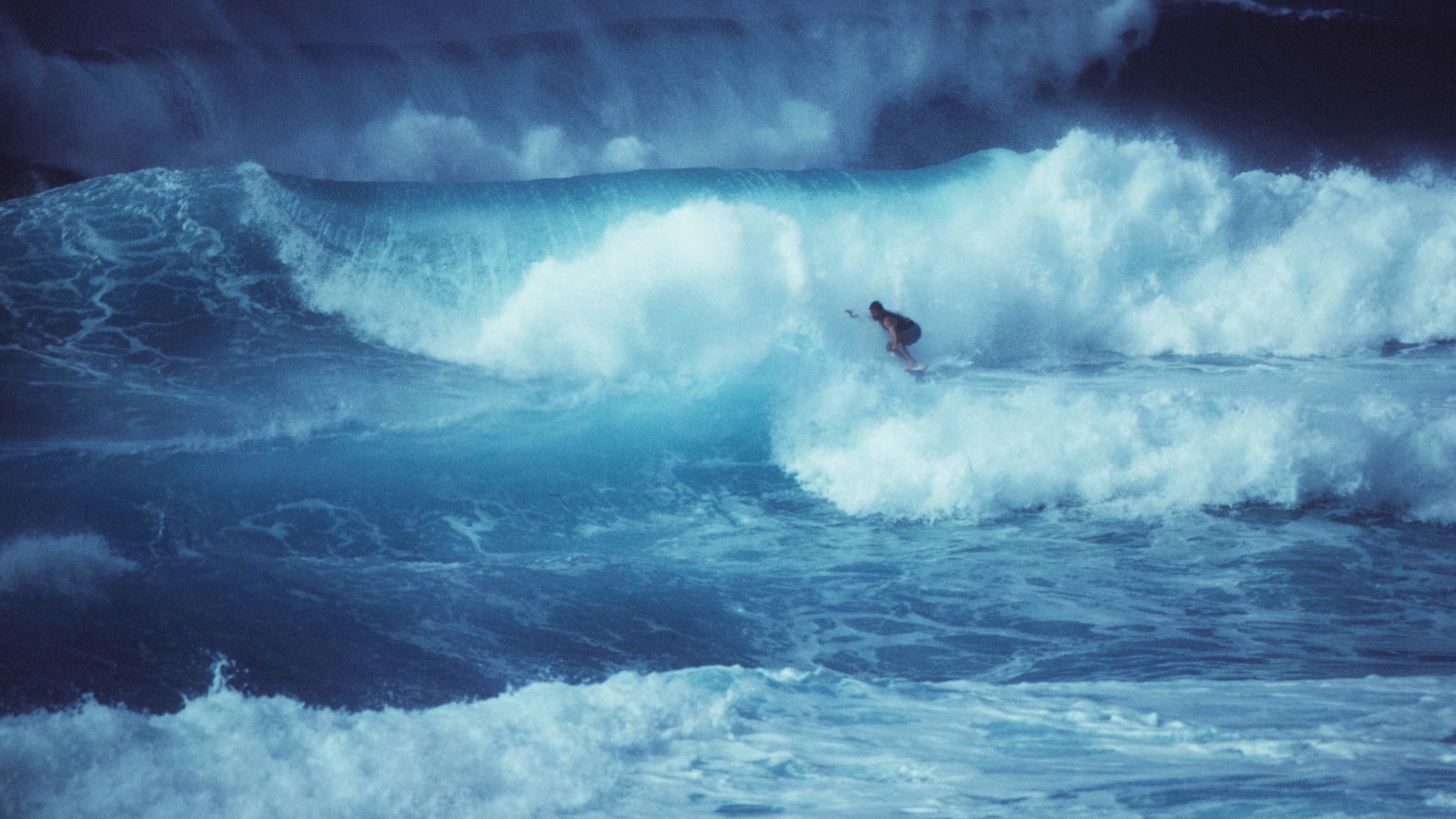 Photo of a surfer riding a large wave in the ocean with cloudy spray, capturing the power and movement of the sea.
