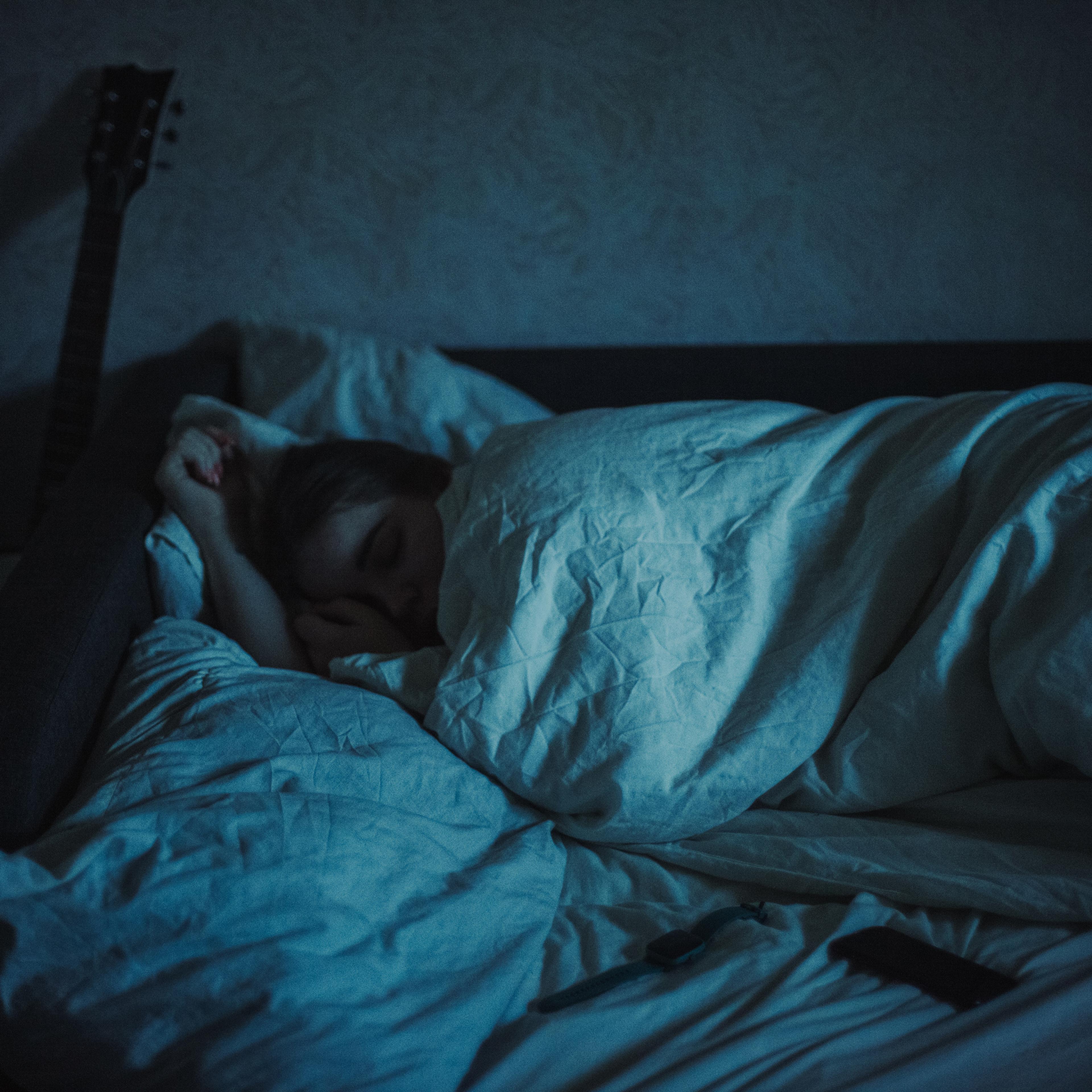 In a dark room a woman is asleep under a duvet and a guitar is leaning against the wall