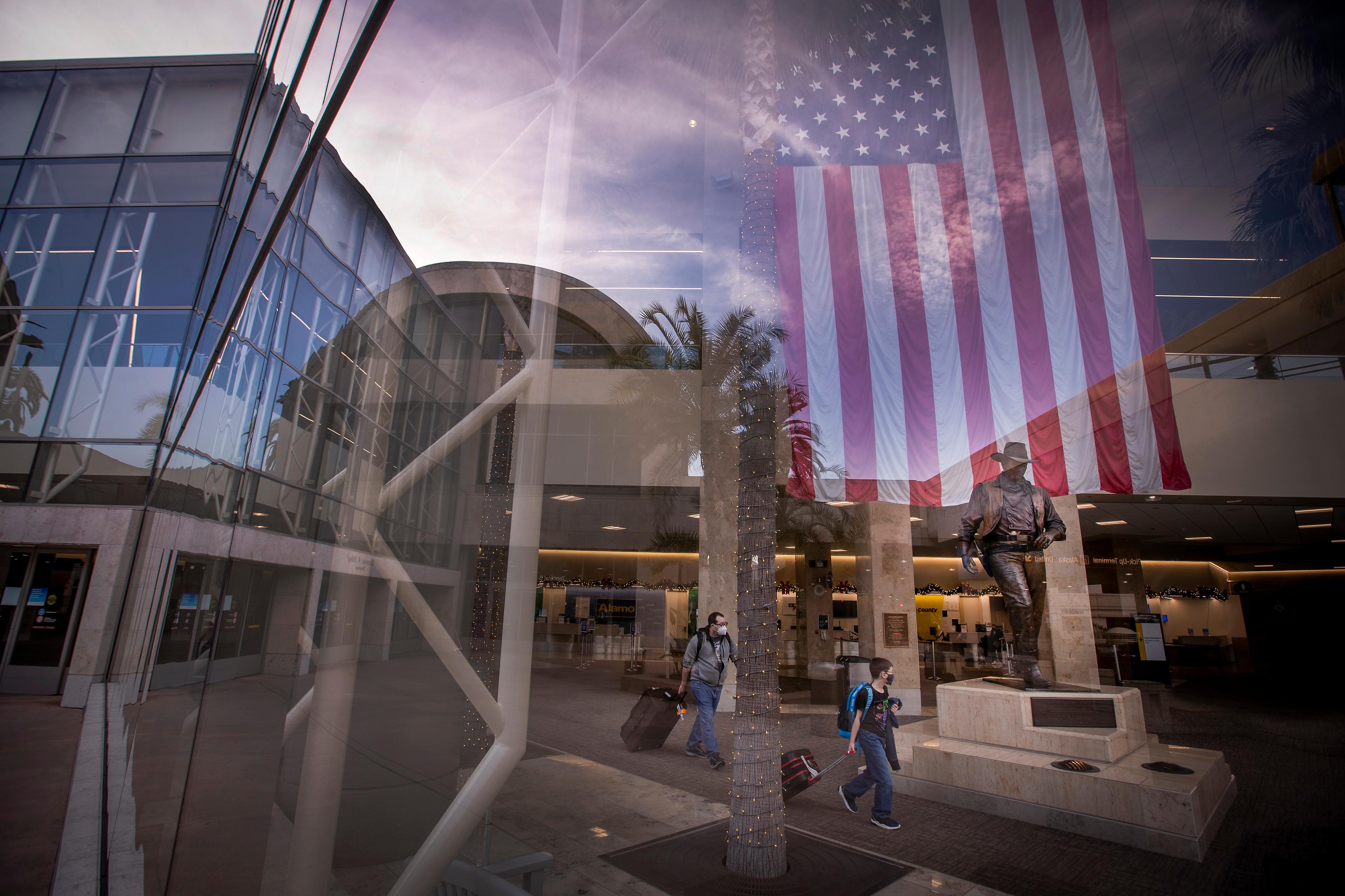 Photo of an airport terminal with travellers, a large US flag and a statue reflected in glass, showing modern architecture.