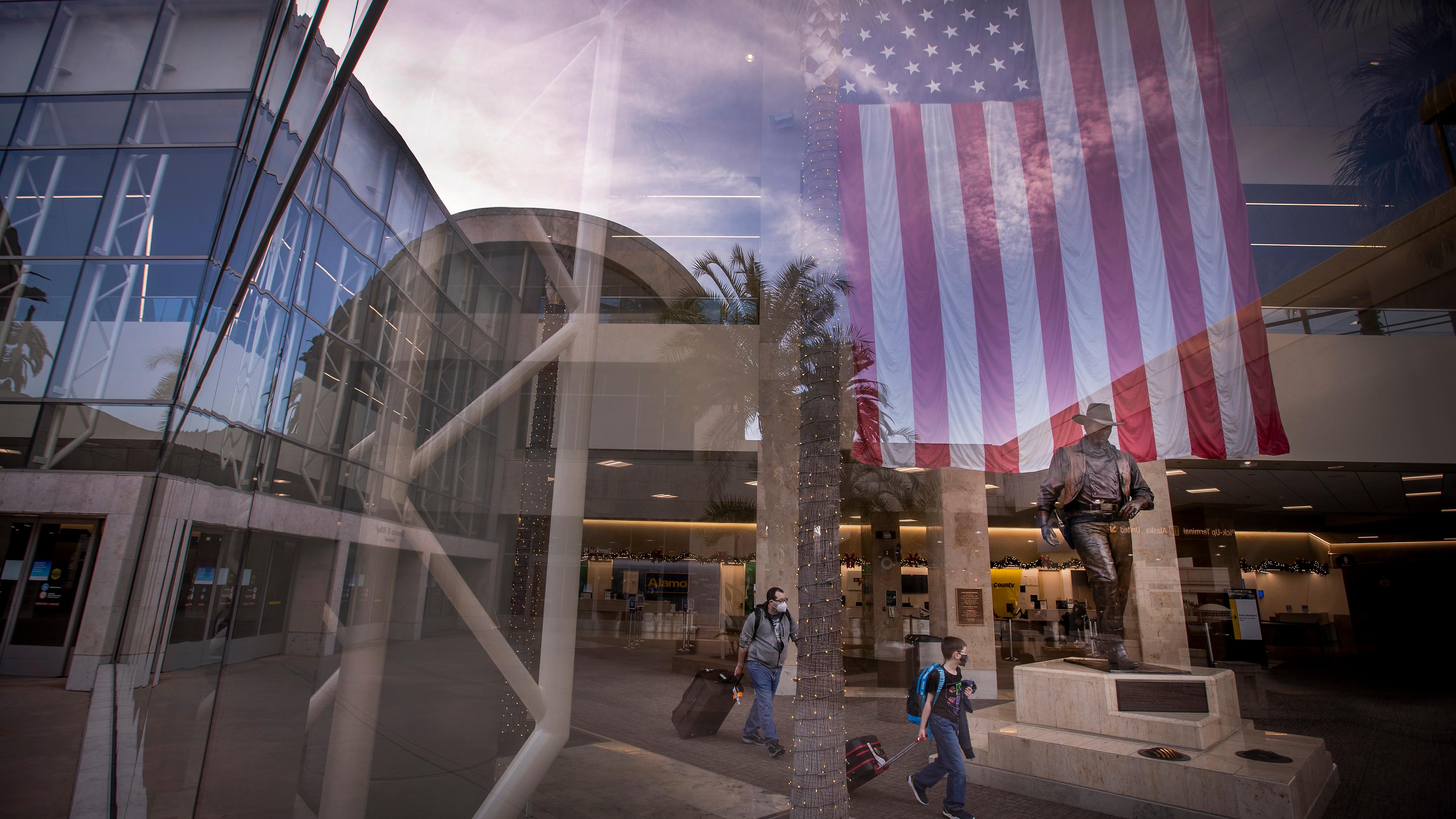 Photo of an airport terminal with travellers, a large US flag and a statue reflected in glass, showing modern architecture.