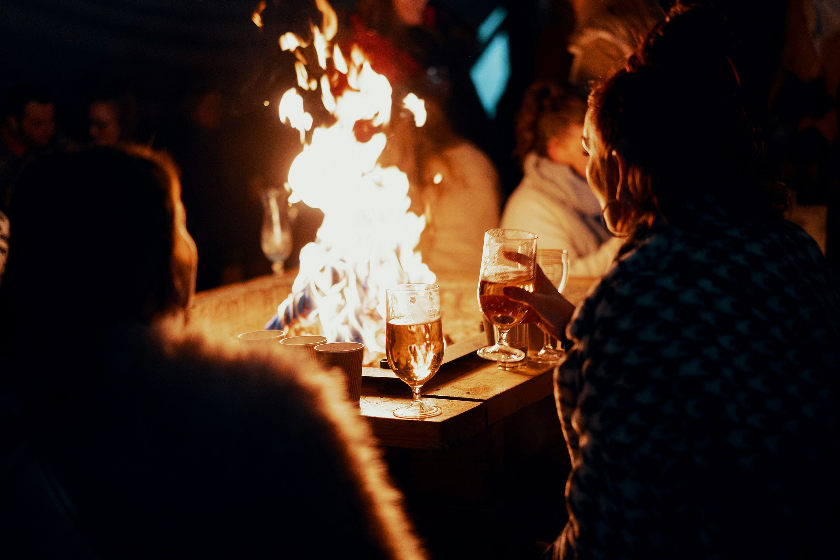 Photo of people around a fire holding drinks, with a warm glow lighting the scene at night.