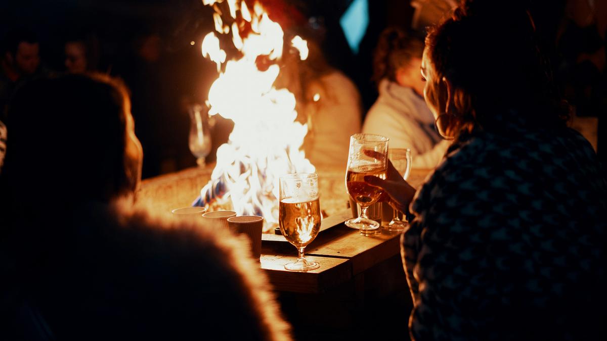 Photo of people around a fire holding drinks, with a warm glow lighting the scene at night.